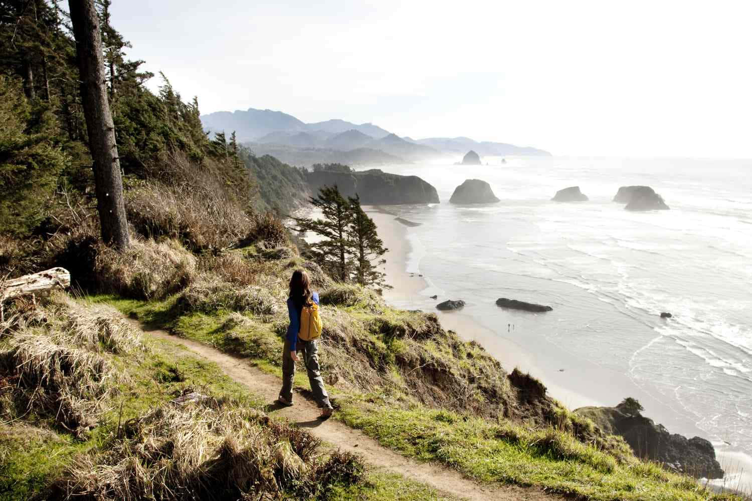Oregon Coast 桌布 壁紙 — 一位背著黃色背包的遠足者沿著一條草叢生的懸崖小徑行走，俯瞰著霧氣瀰漫的海洋、岩石海岸和遠處的山脈。