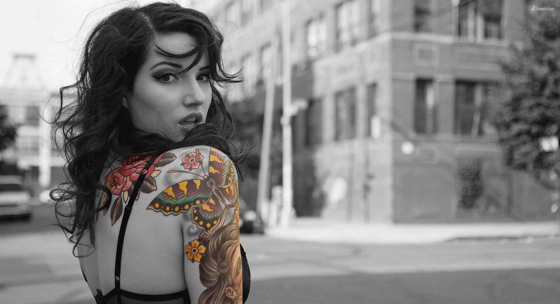 Tattoo 桌布 壁紙 — 一位背部有鮮豔花卉和蝴蝶紋身的女性，在模糊的城市街道上回頭望向肩膀後方。