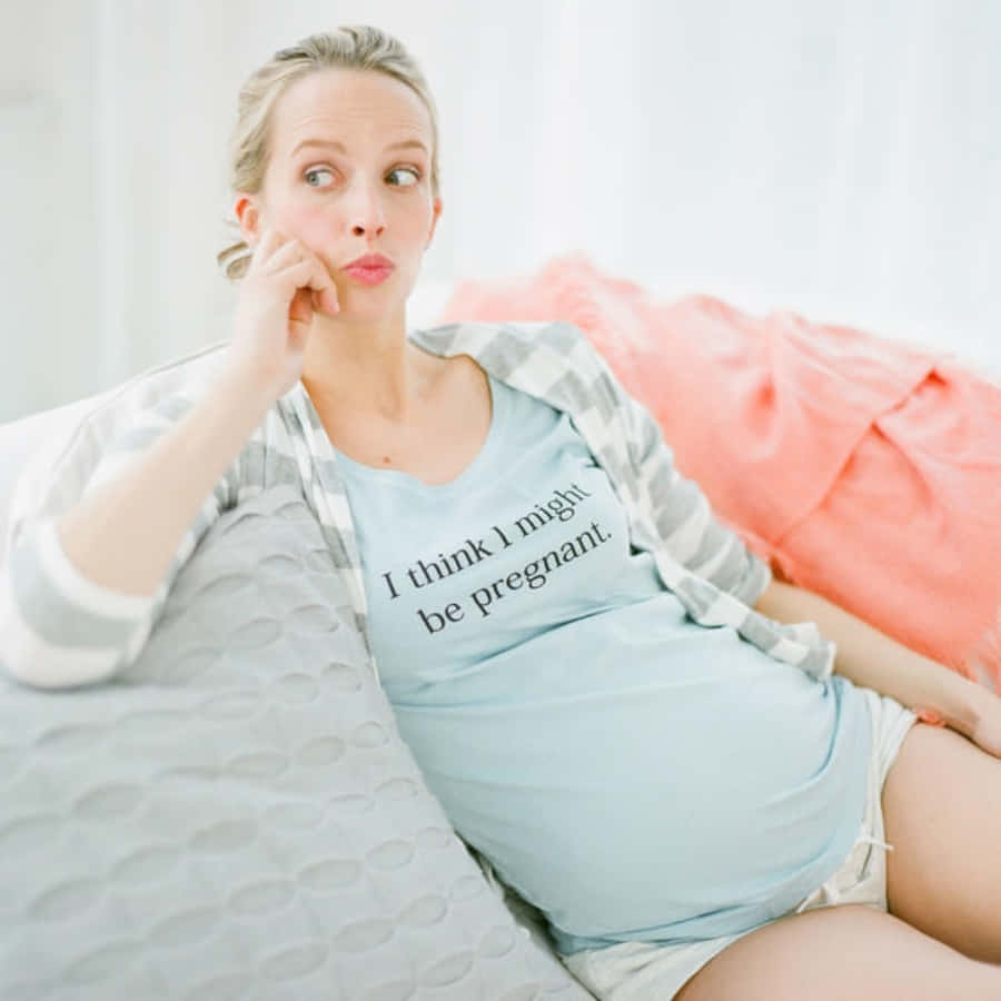 Pregnant 桌布 壁紙 — 一位腹部微圓的女性坐在沙發上，穿著一件寫著「我想我可能懷孕了」的襯衫，若有所思地看向一旁。