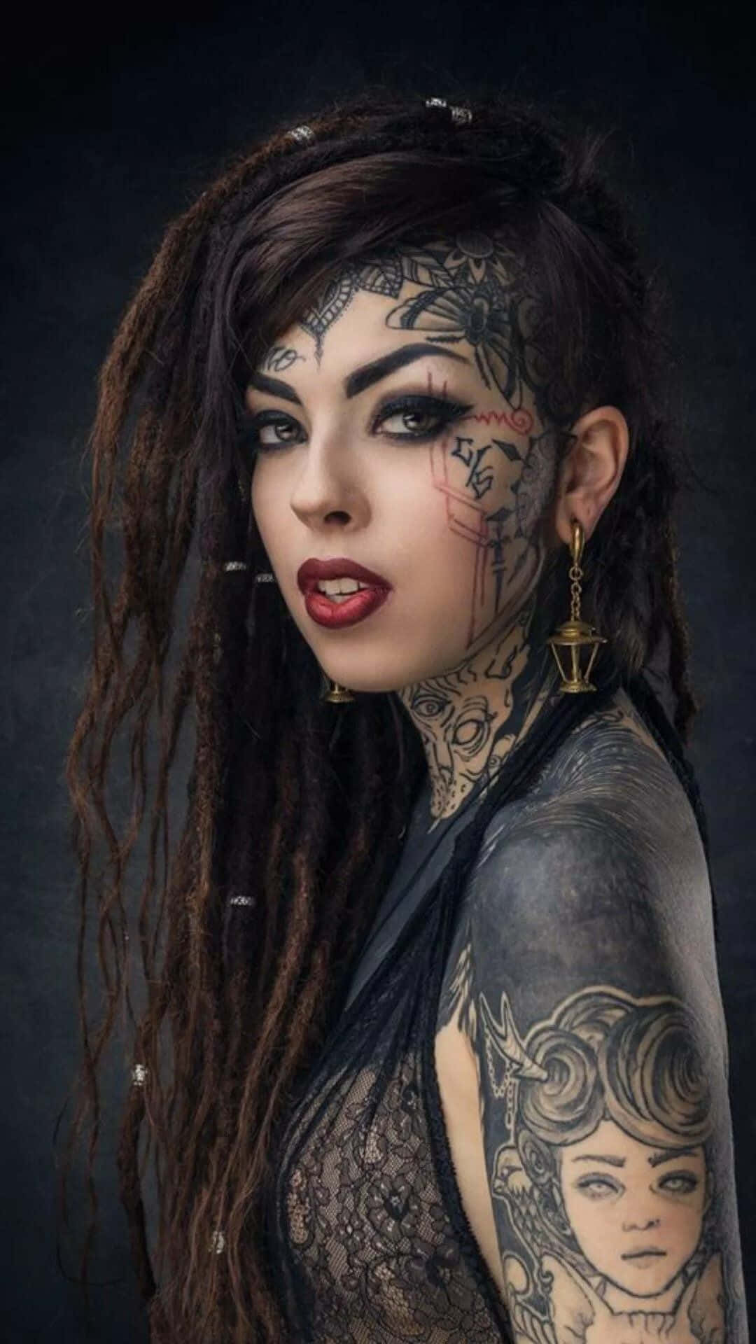 Tattoo Girl 桌布 壁紙 — 一位臉上、頸部和手臂上有複雜紋身的女性，在黑暗陰鬱的背景中強烈凝視。