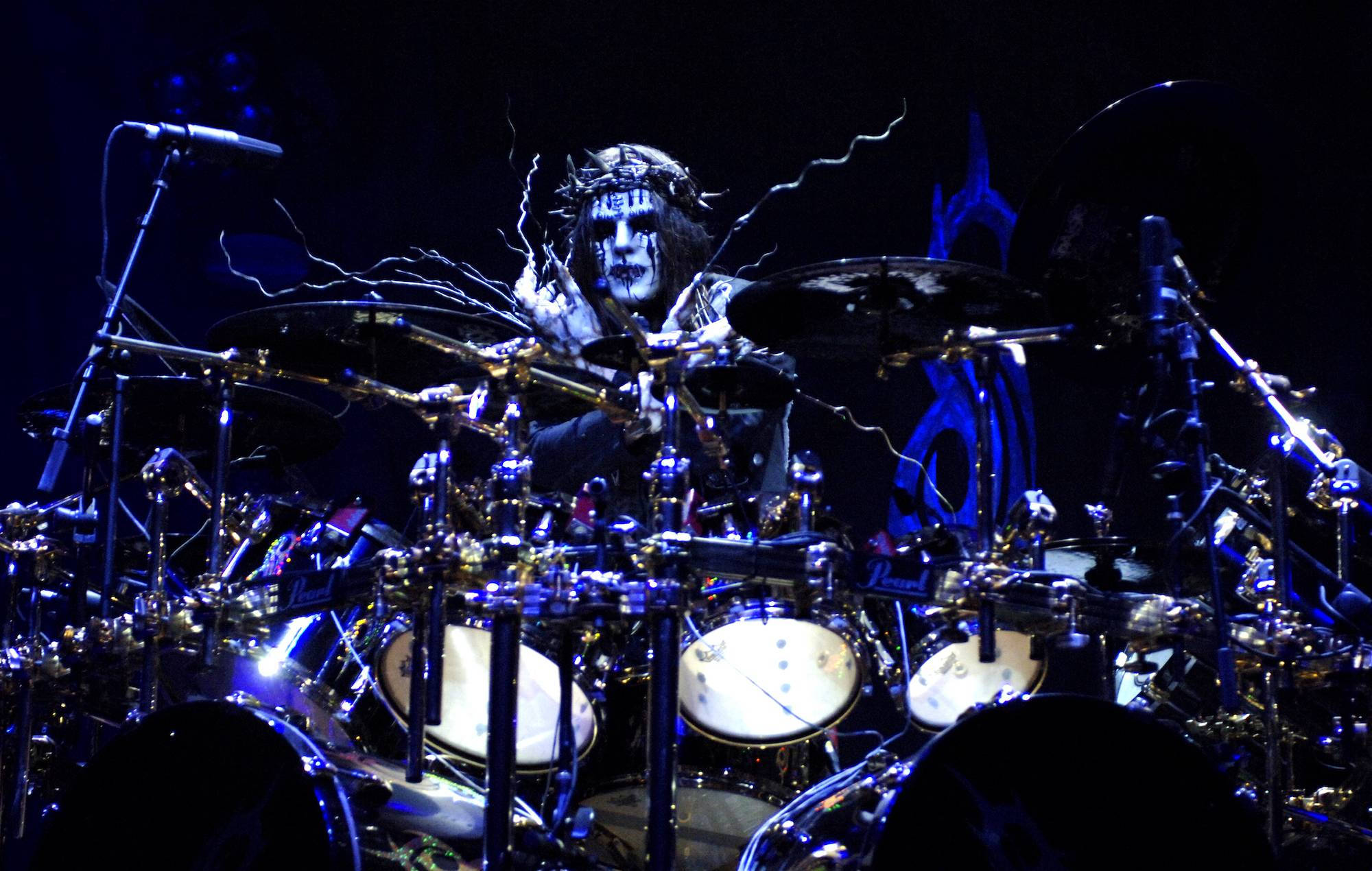 Joey Jordison 桌布 壁紙 — 一位臉上塗著白色顏料、戴著皇冠的鼓手，在藍色舞台燈光下演奏閃閃發光的鼓組。