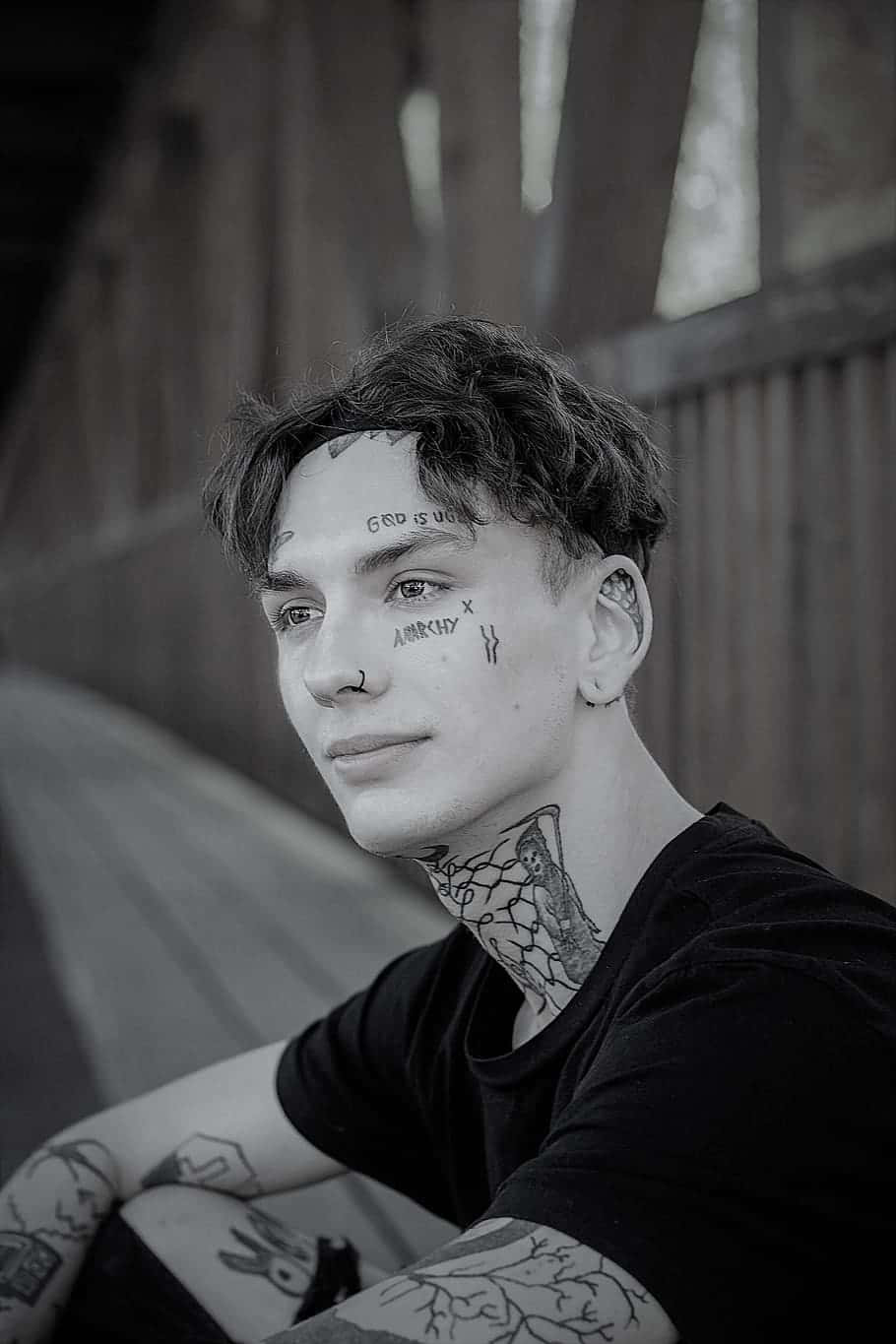 Tattoo 桌布 壁紙 — 一位臉上帶有刺青和文字的年輕男子在昏暗的木結構中平靜地凝視。