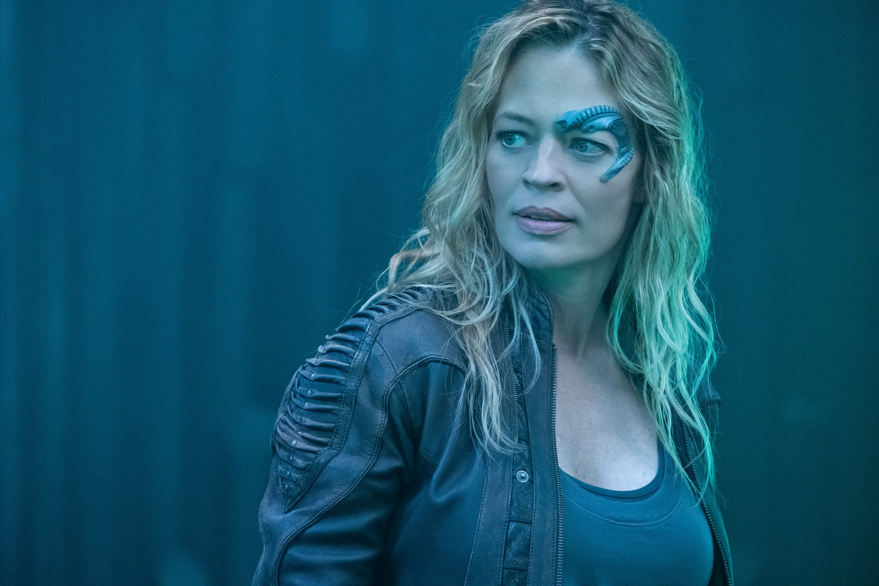 Jeri Ryan 桌布 壁紙 — 一位臉上帶有發光藍色標記、波浪金髮的女性，穿著皮夾克，背景為青綠色。