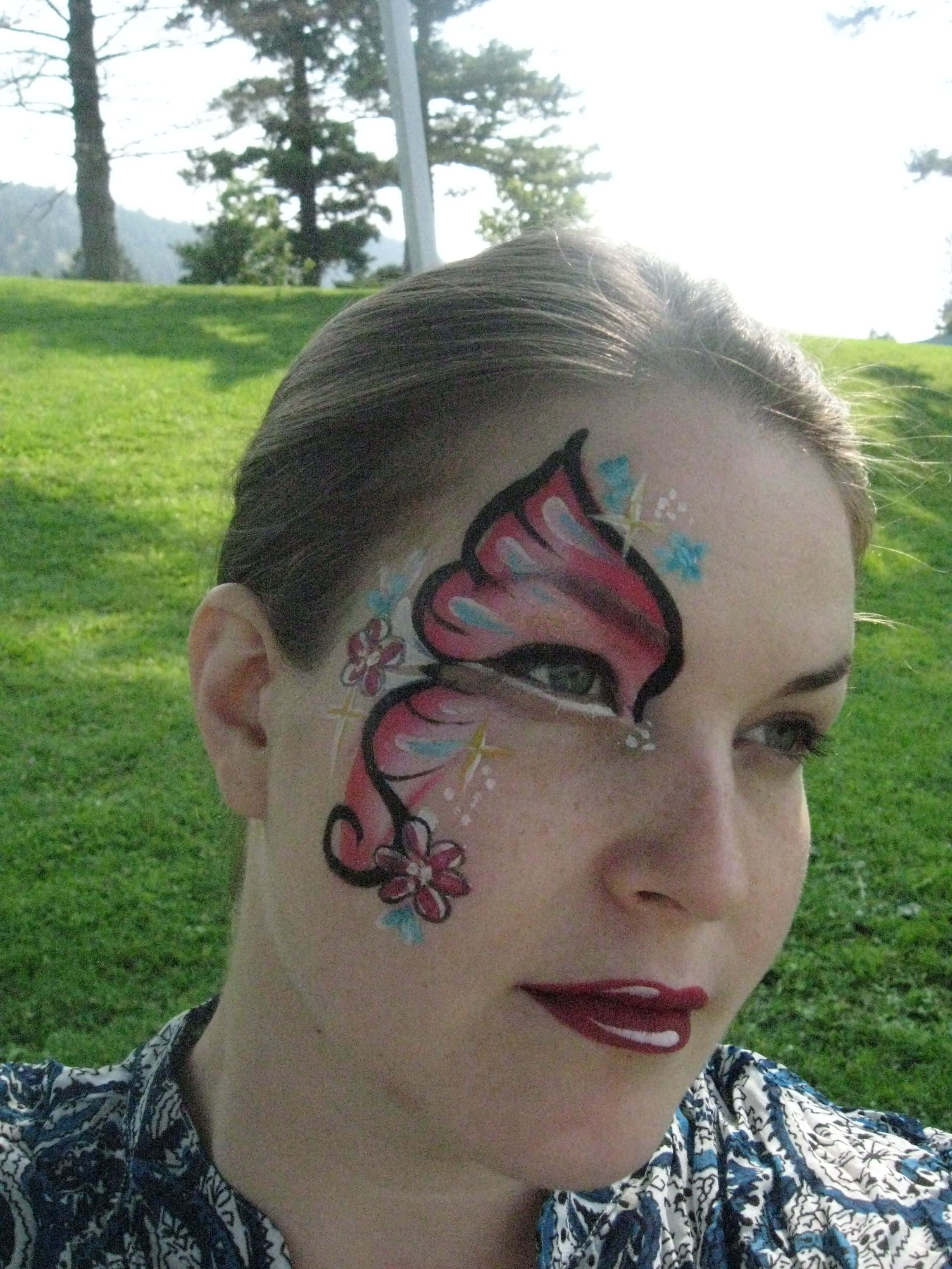 Butterfly Face Painting 桌布 壁紙 — 一位臉上畫有鮮豔蝴蝶彩繪的女性，在陽光明媚的日子裡，以草地山坡和樹木為背景，在戶外微笑。