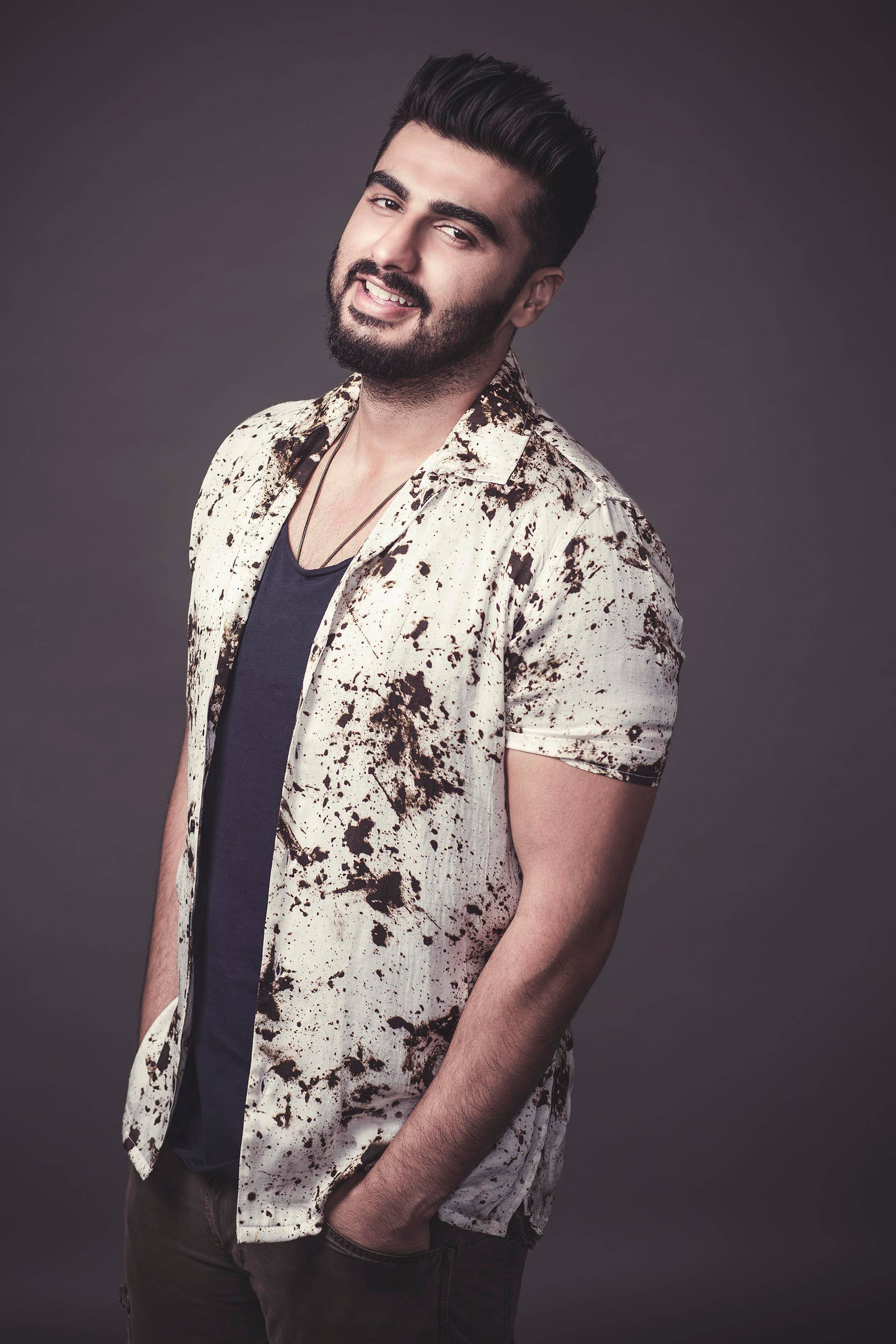 Arjun Kapoor 桌布 壁紙 — 一位自信滿滿、留著鬍子、頭髮造型時尚的男士，在柔和的灰色背景前，身穿深色T恤外搭一件濺有污漬的白襯衫，面帶微笑。