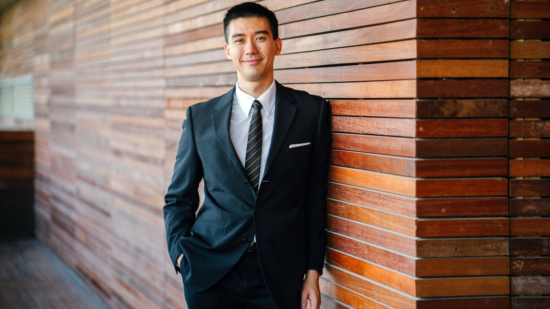 suit 桌布 壁紙 — 一位自信滿滿、身穿俐落西裝的男子，輕鬆地靠著暖色調的木條牆，露出溫柔的微笑。