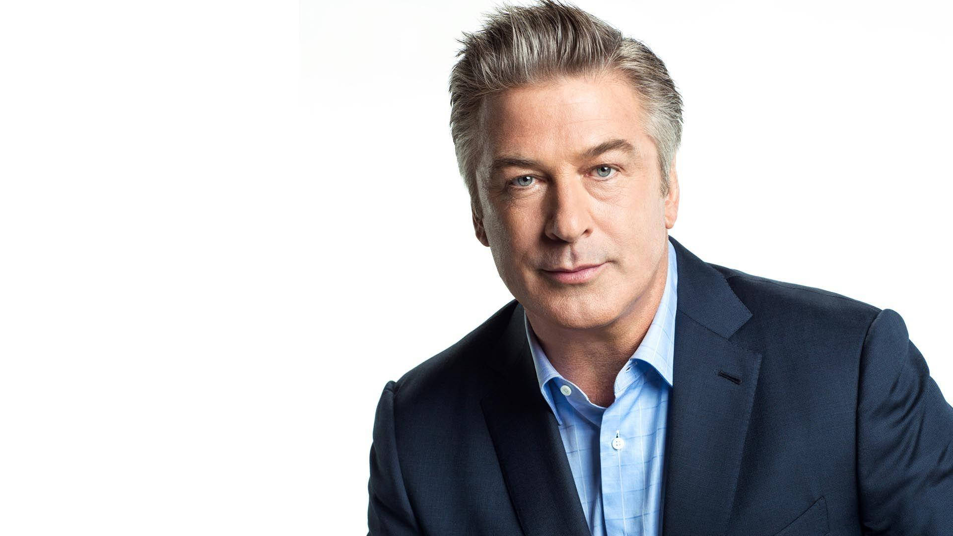 Alec Baldwin 桌布 壁紙 — 一位自信的中年男士，灰白頭髮，身穿深色西裝和淺藍色襯衫，背景是純白色。