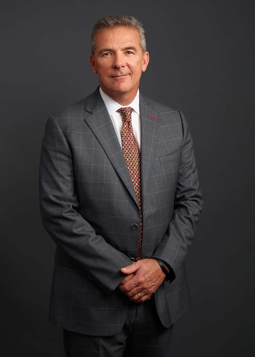 Urban Meyer 桌布 壁紙 — 一位自信的中年男士身穿灰色格紋西裝和圖案領帶，雙手交握，站在深灰色攝影棚背景前。