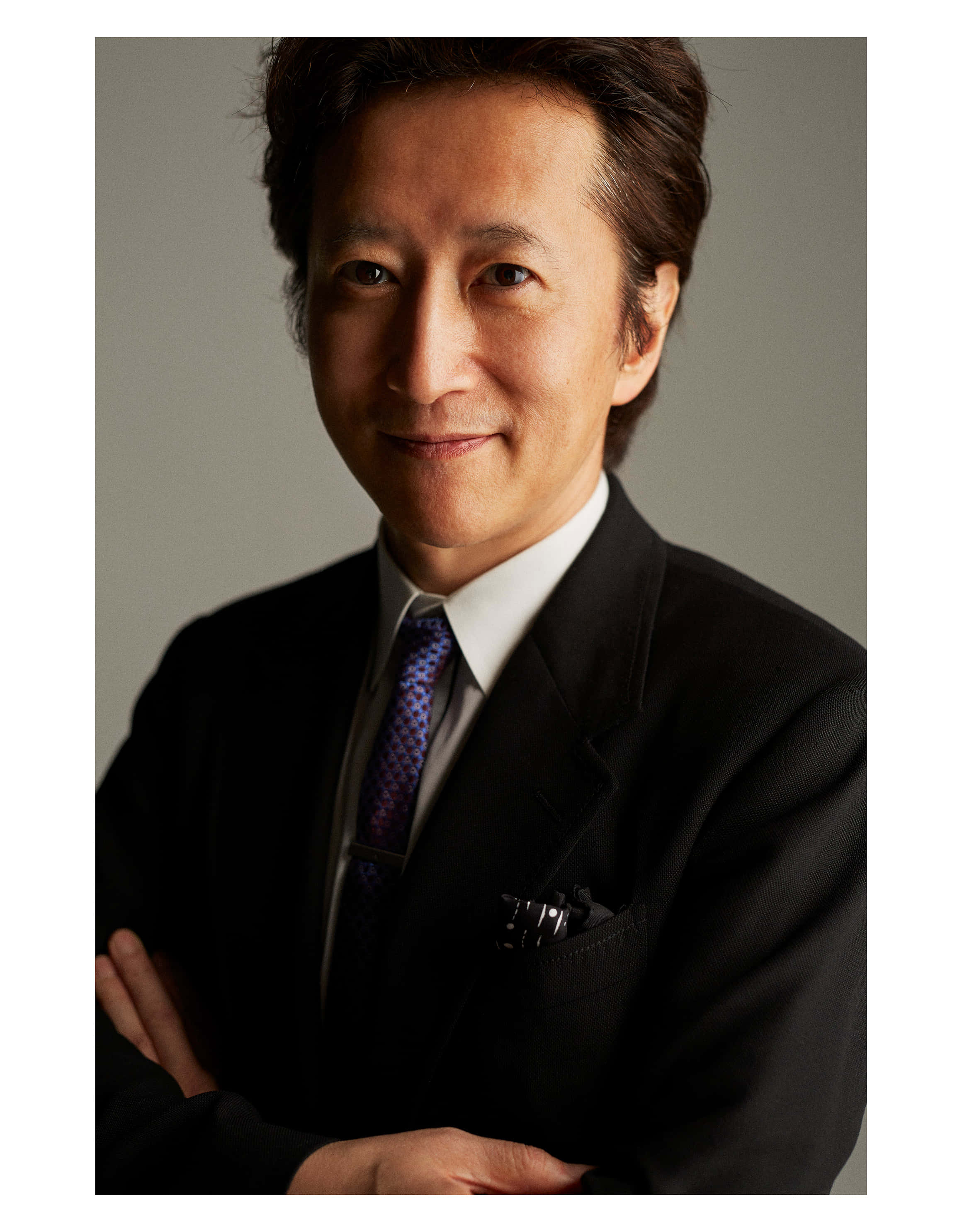 Hirohiko Araki 桌布 壁紙 — 一位自信的亞洲男士身穿黑色西裝和圖案領帶，雙臂交叉，站在中性灰色背景前。