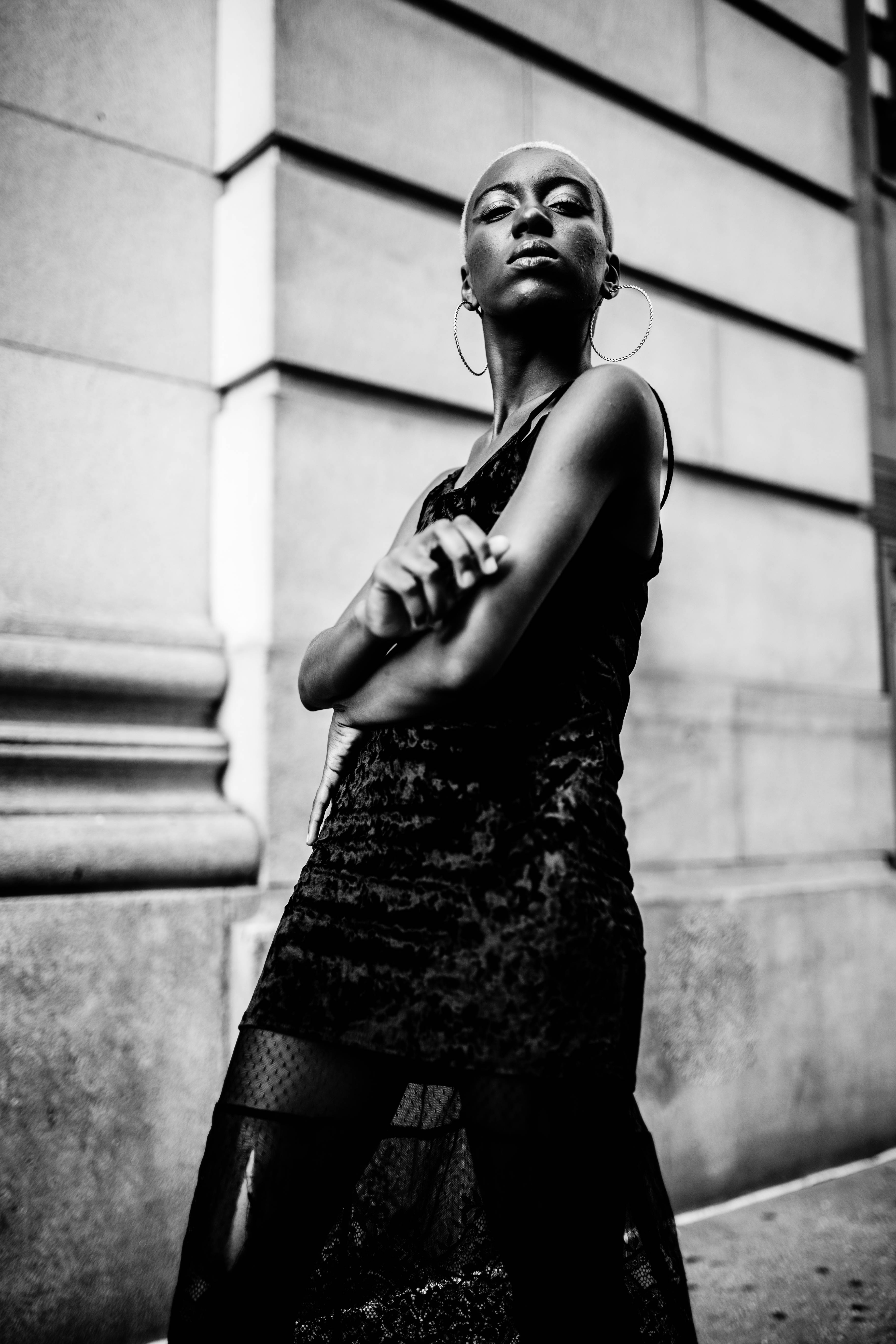 Black Girl 桌布 壁紙 — 一位自信的光頭女性穿著蕾絲連衣裙，雙臂交叉靠在石頭建築上，形成一幅引人注目的黑白城市肖像。
