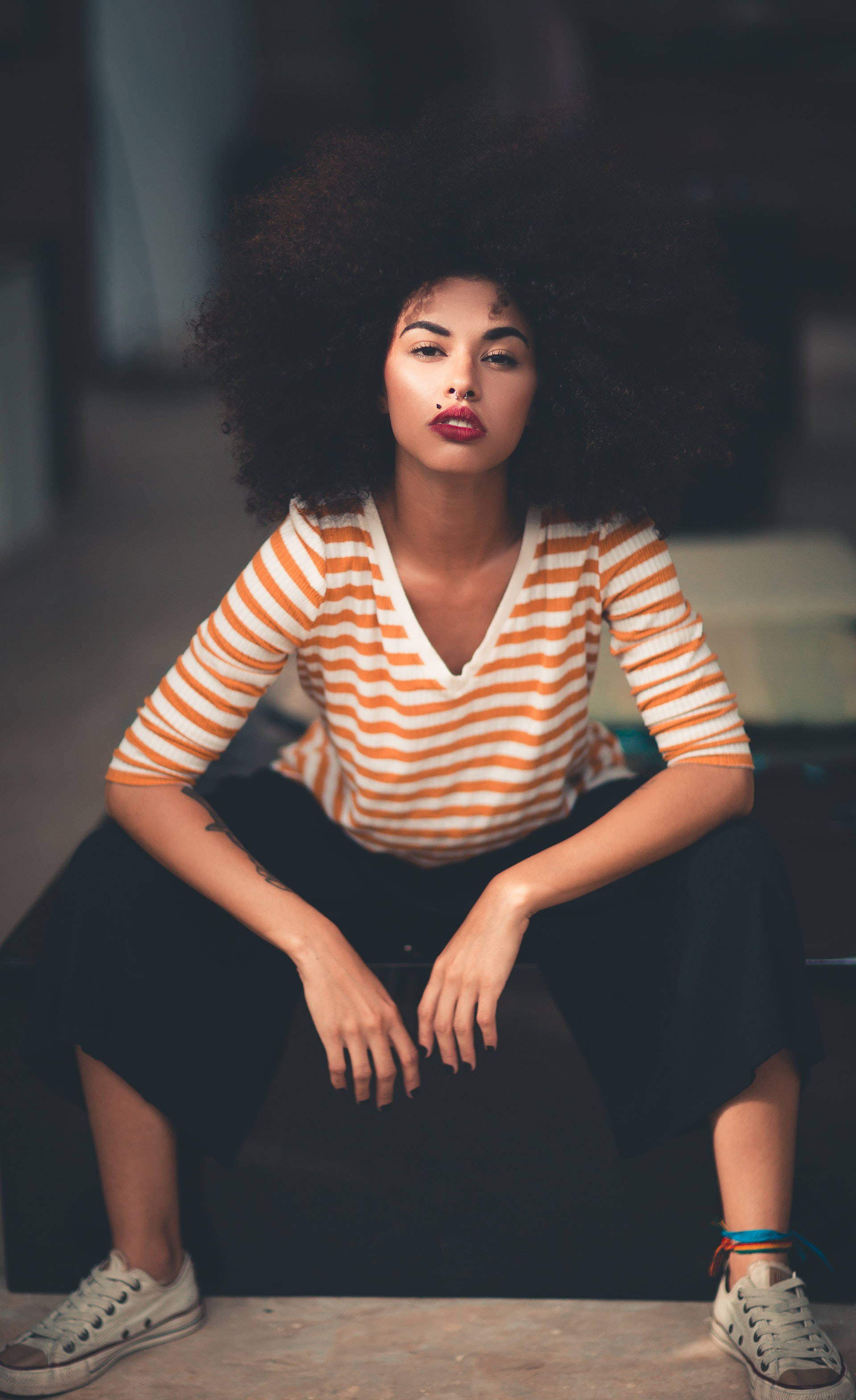 Black Girl 桌布 壁紙 — 一位自信的女人，留著大爆炸頭，盤腿坐著，穿著橘白條紋襯衫和運動鞋。