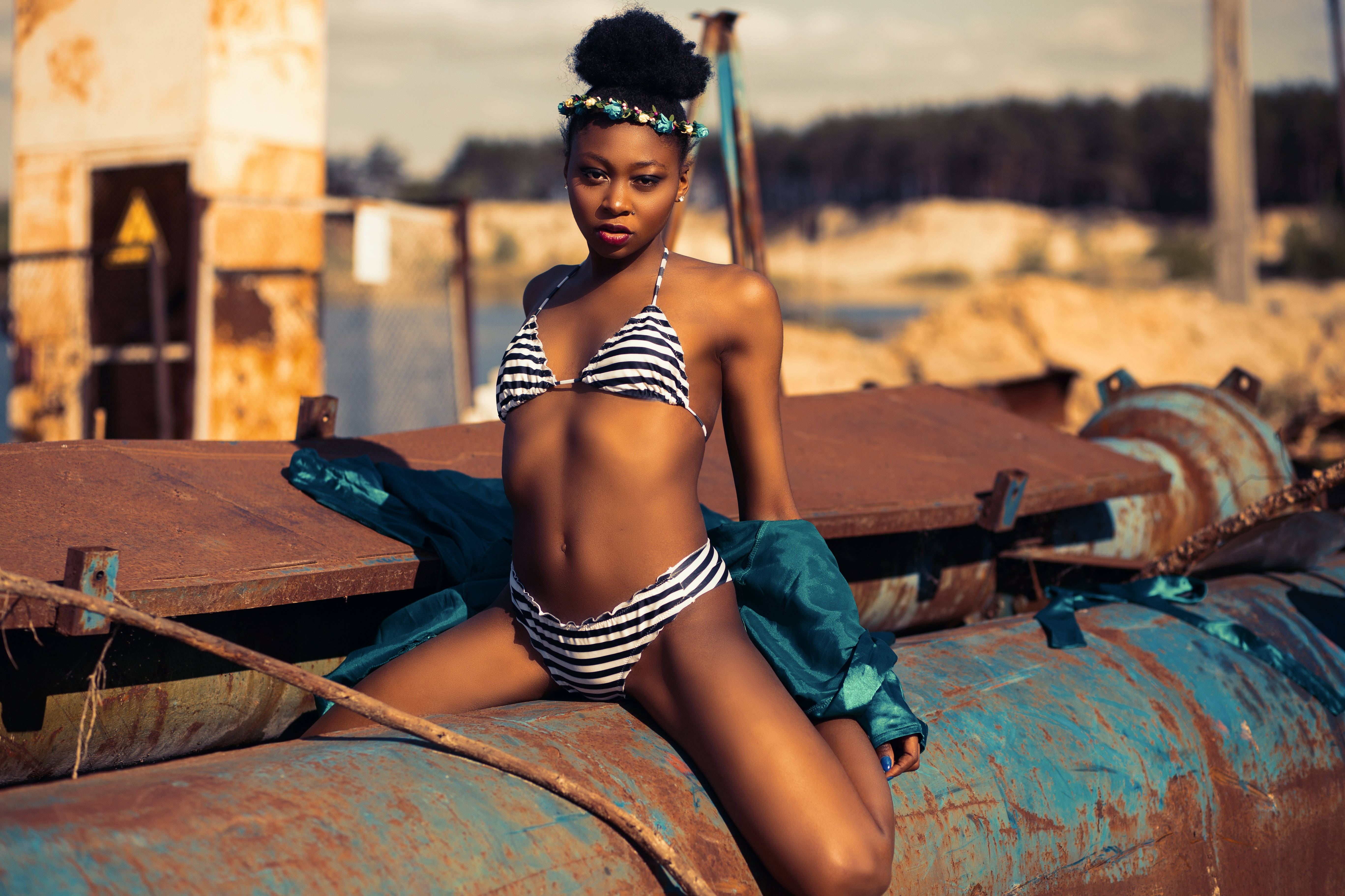 bikini model 桌布 壁紙 — 一位自信的女人穿著條紋比基尼，坐在陽光明媚的廢棄庭院中生鏽的工業管道上。