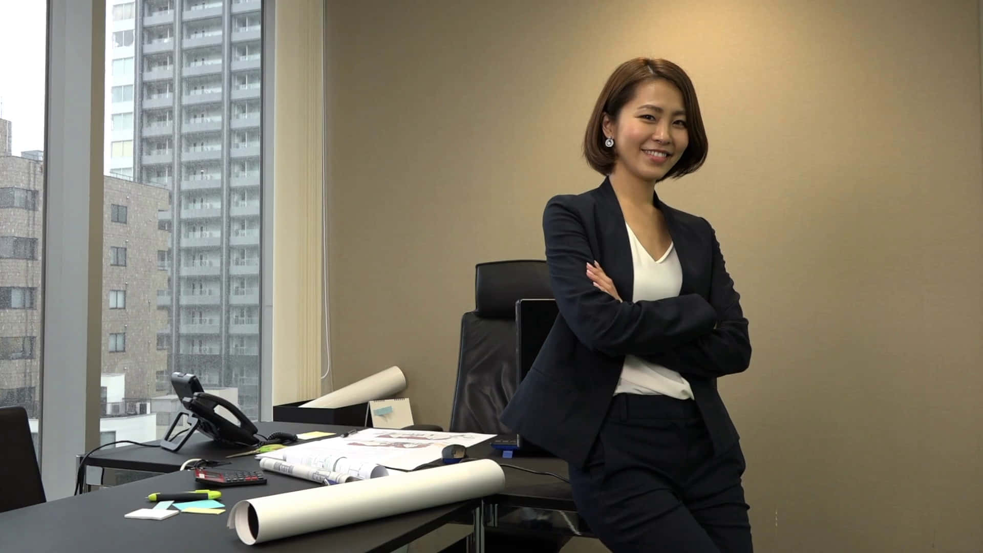 Woman 桌布 壁紙 — 一位自信的女商人穿著深色西裝，在現代辦公室裡交叉雙臂站立，微笑對著鏡頭，身後窗戶可見城市建築。