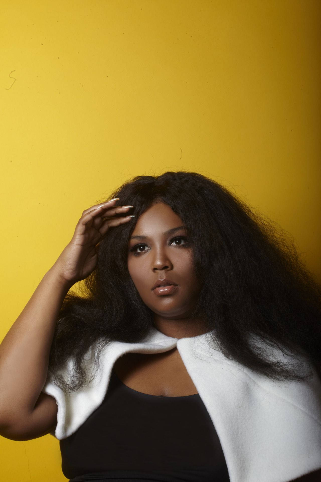 Lizzo 桌布 壁紙 — 一位自信的女性，擁有豐盈的黑髮，在鮮黃色背景前擺姿勢，手輕輕觸碰額頭。
