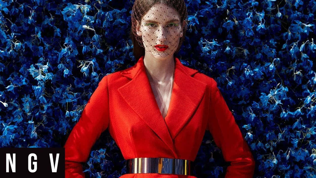 Red Coat 桌布 壁紙 — 一位自信的女性穿著大膽的紅色西裝外套和金屬腰帶，站在密集的鮮藍色花朵背景前。