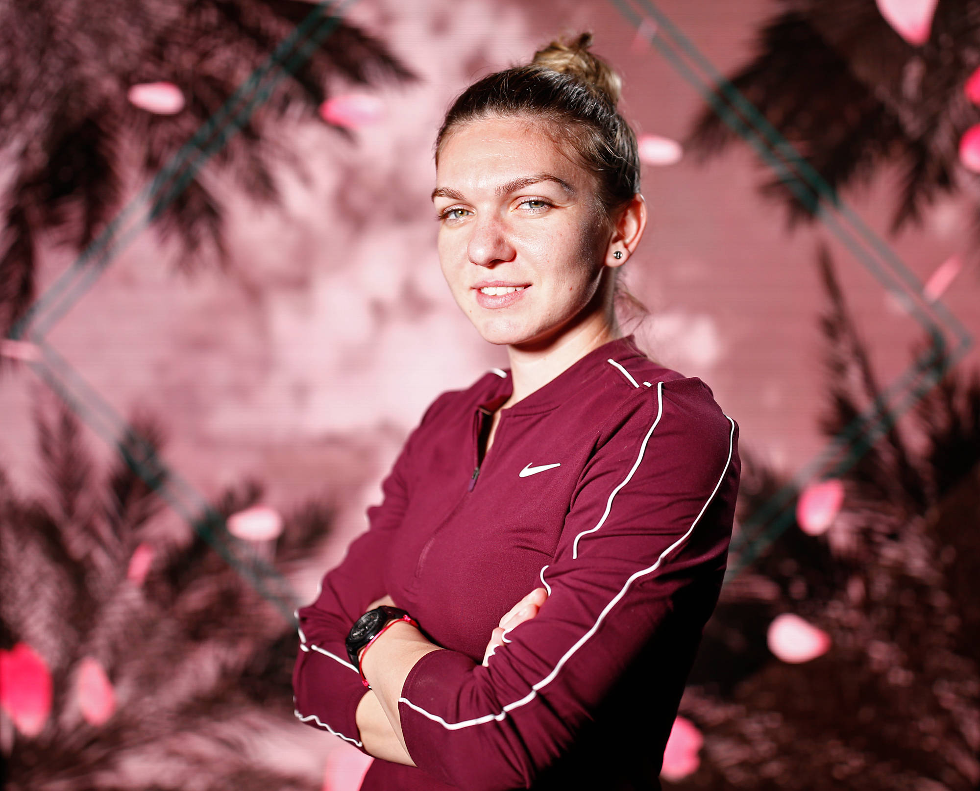 運動 桌布 壁紙 — 一位自信的女性穿著栗色運動服，在粉紅色棕櫚葉和飄落花瓣的背景前交叉雙臂站立。