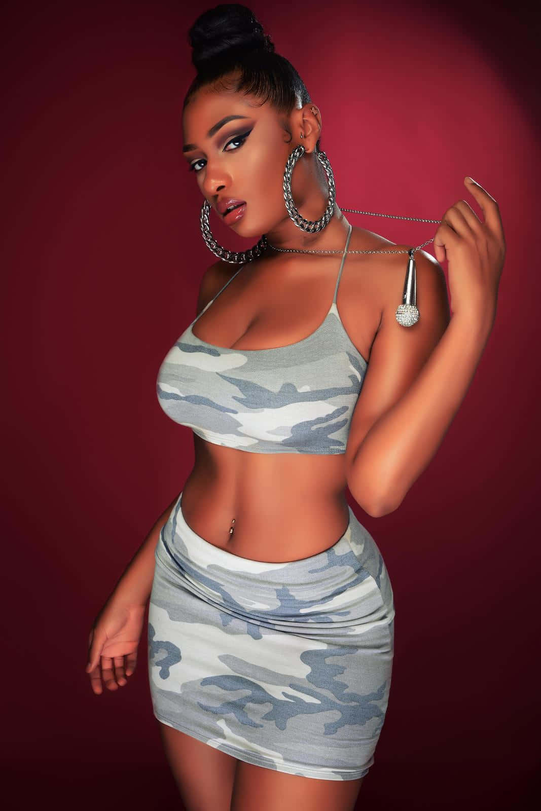Megan Thee Stallion 桌布 壁紙 — 一位自信的女性穿著迷彩短上衣和迷你裙，在深紅色工作室背景前戴著超大耳環擺姿勢。