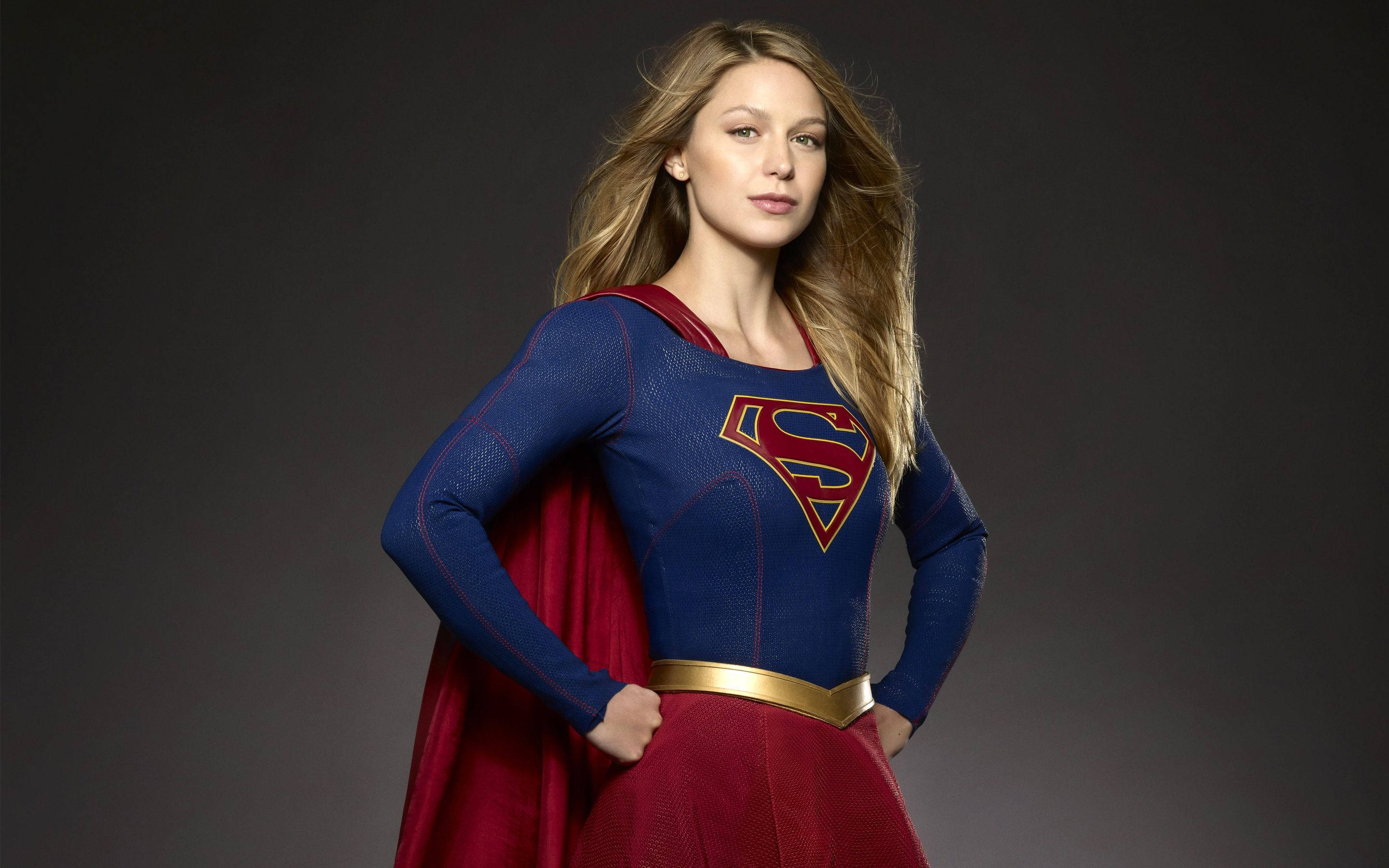 Superwoman 桌布 壁紙 — 一位自信的女性超級英雄，身穿藍色和紅色服裝，配金色腰帶，站在深灰色工作室背景前。