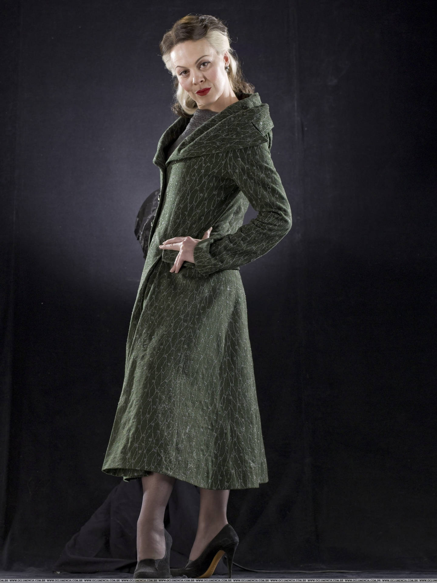 Slytherin 桌布 壁紙 — 一位自信的女性身穿質感綠色外套，一手叉腰，在深色背景前自信站立。