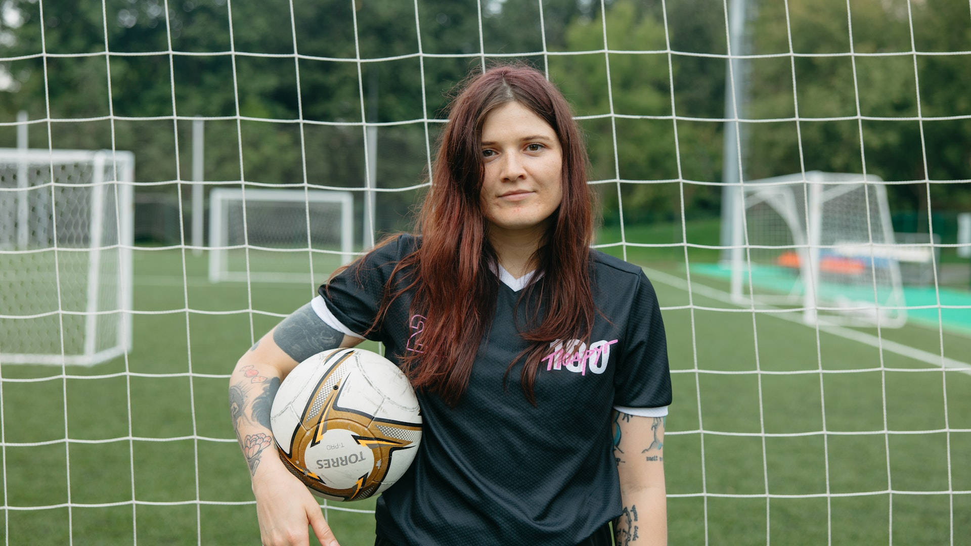 Athletic 桌布 壁紙 — 一位自信的女足球員帶有刺青，在綠色球場上於球門網前拿著一個球。