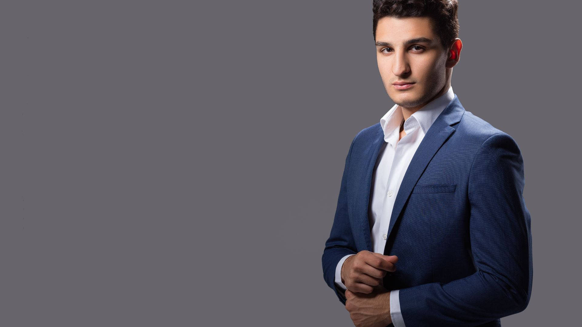 suit 桌布 壁紙 — 一位自信的年輕男子穿著俐落的藍色西裝，站在純灰色攝影棚背景前，直視鏡頭。