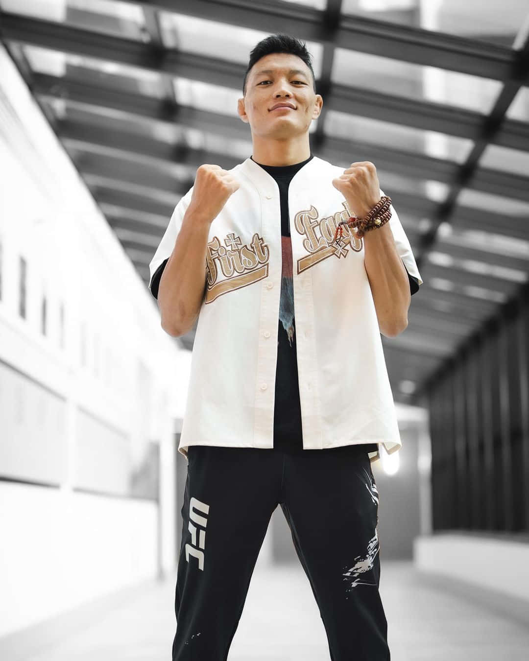 UFC 桌布 壁紙 — 一位自信的戰士身穿白色棒球衫和黑色UFC褲，在現代玻璃牆走廊中舉拳擺姿勢。
