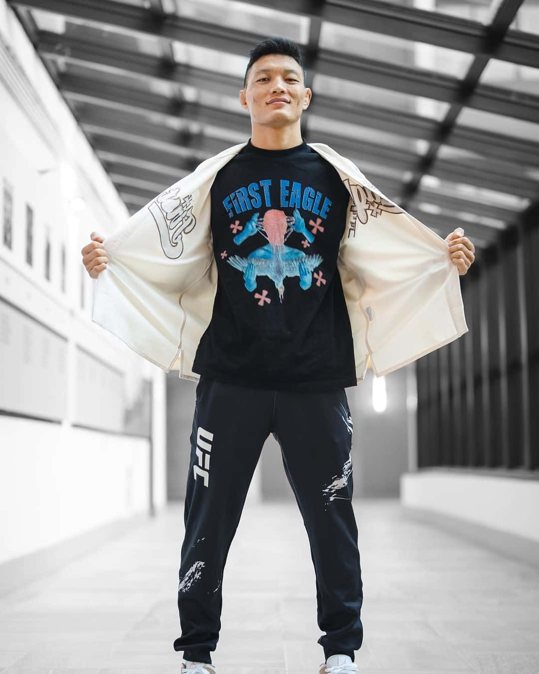 Celebrity 桌布 壁紙 — 一位自信的拳手身穿UFC裝備，站在現代走廊中，拉開奶油色外套，露出圖案T恤。