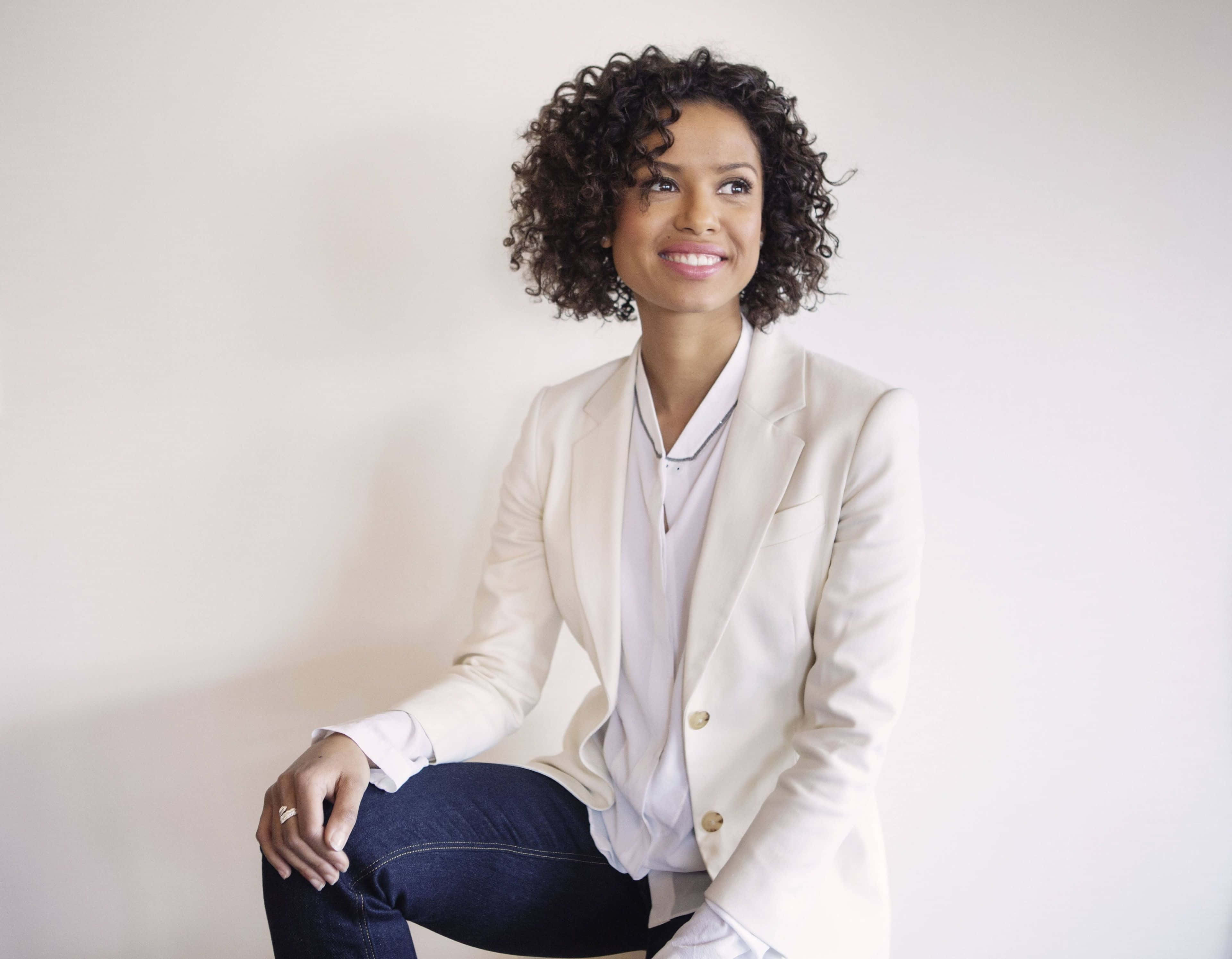 Gugu Mbatha-raw 桌布 壁紙 — 一位自信的捲髮女性，身穿米色西裝外套，坐在純白牆前微笑。