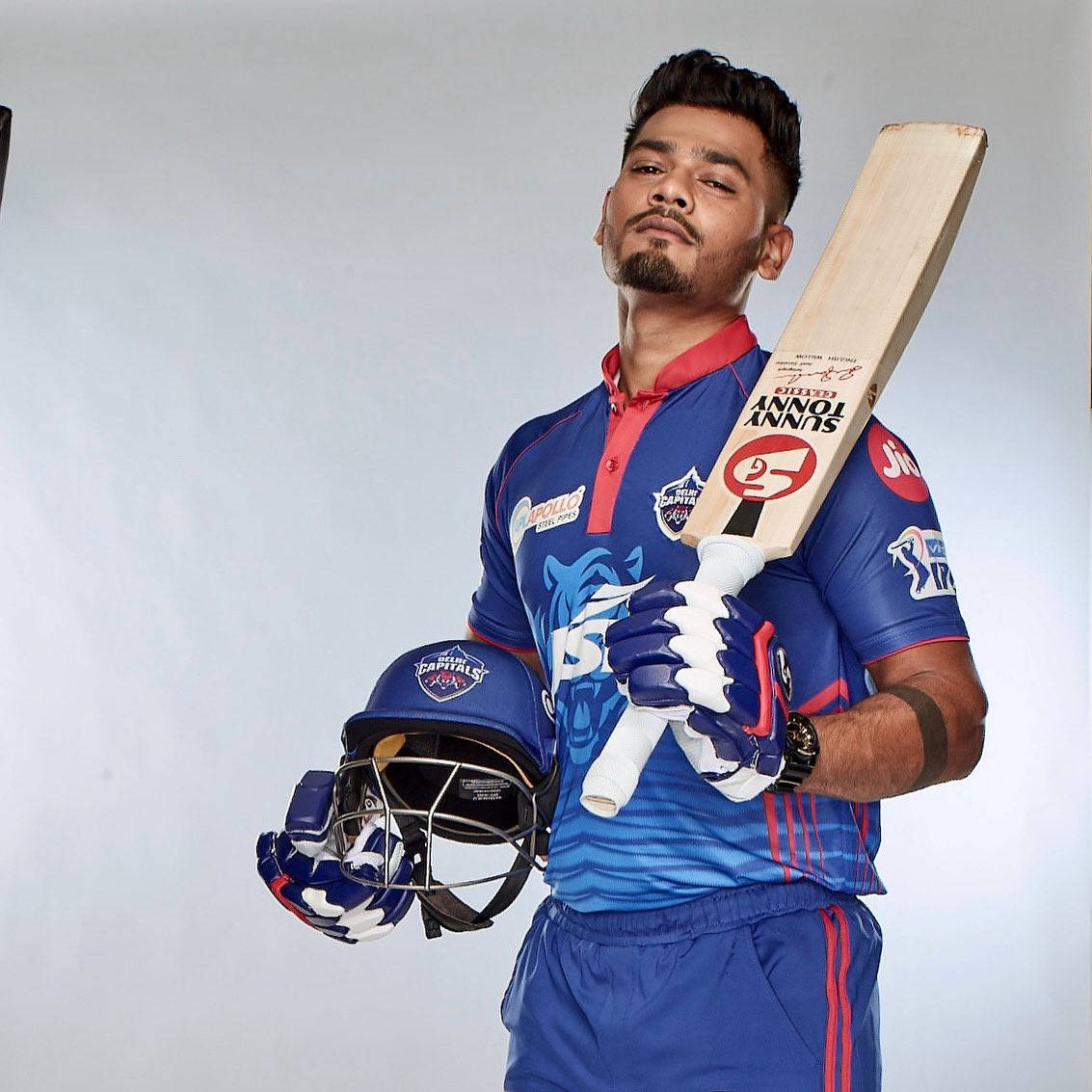 Delhi Capitals 桌布 壁紙 — 一位自信的板球運動員身穿藍色德里首都隊球衣，在純色工作室背景前手持球棒和頭盔。
