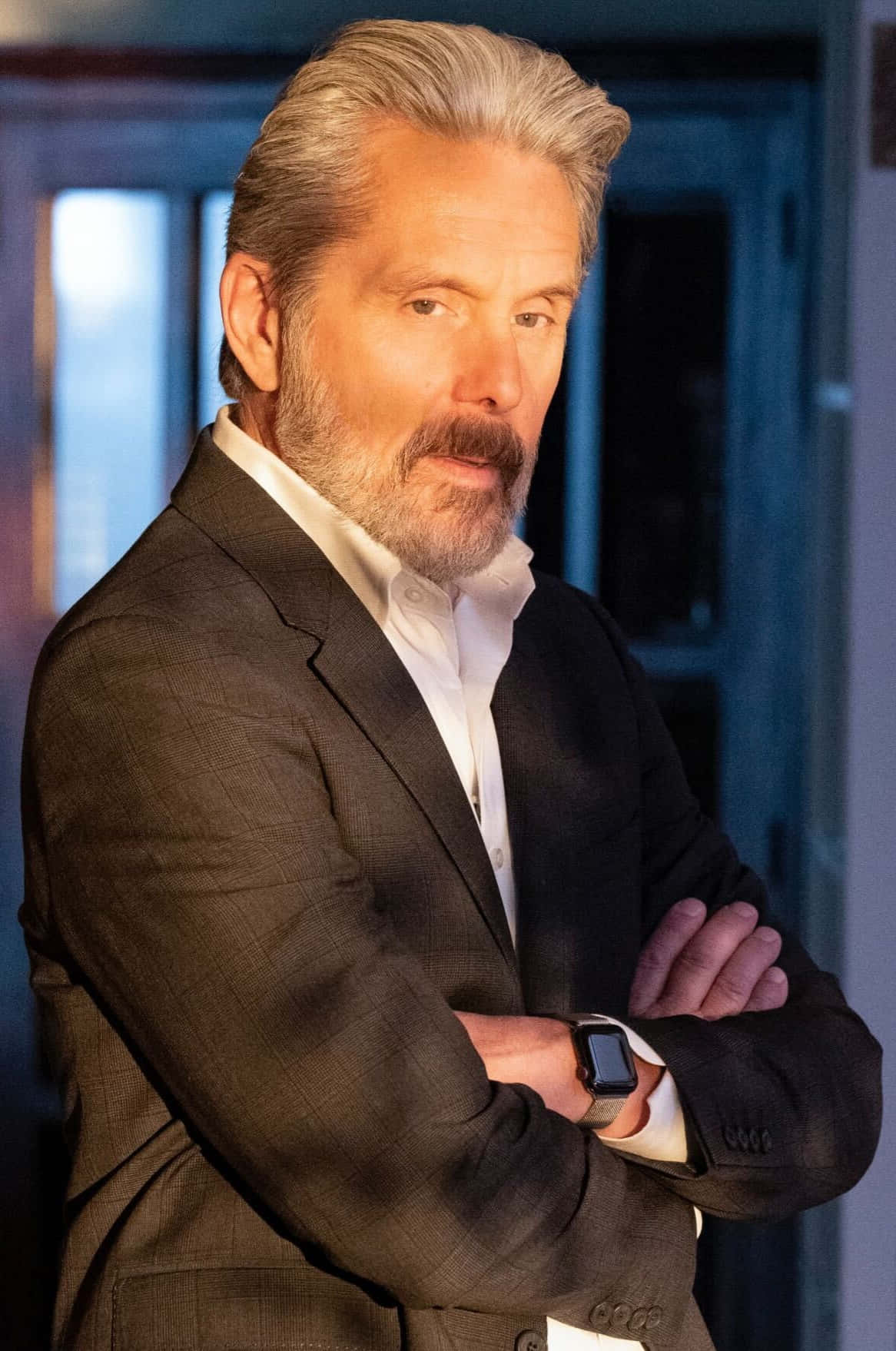 Gary Cole 桌布 壁紙 — 一位自信的灰髮男人穿著深色西裝，在昏暗的現代室內交叉雙臂站著。