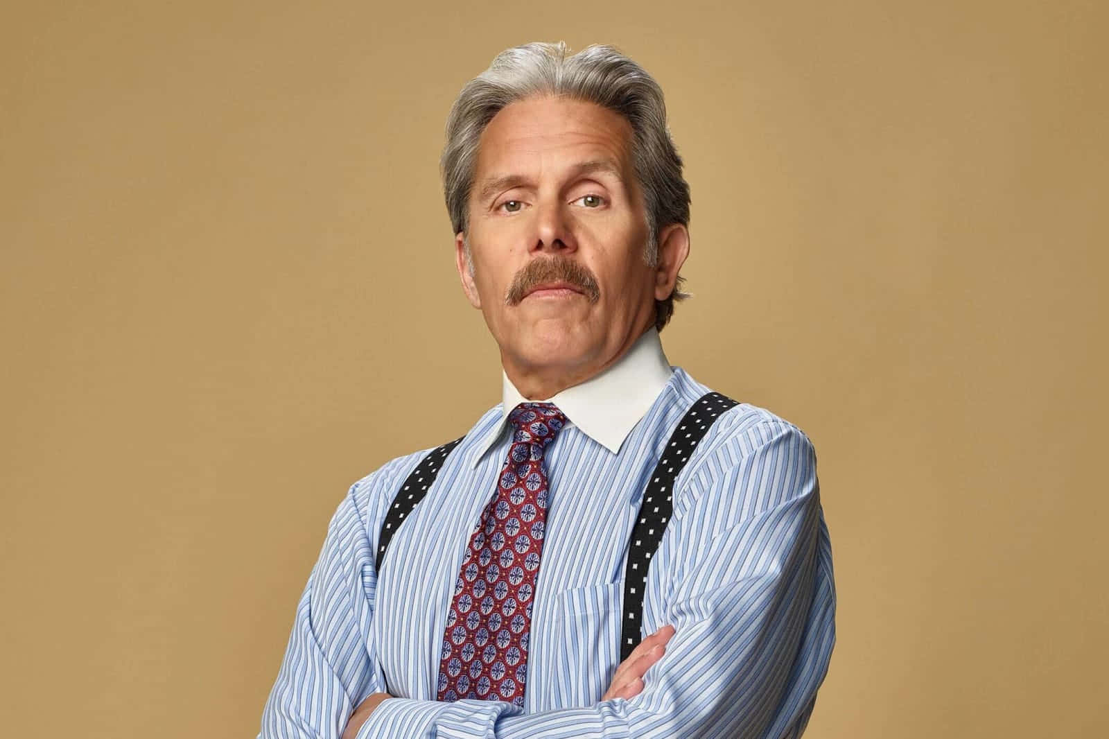 Gary Cole 桌布 壁紙 — 一位自信的灰髮鬍鬚男人，雙手交叉，身穿條紋襯衫、吊帶褲帶和圖案領帶，站在米色背景前。