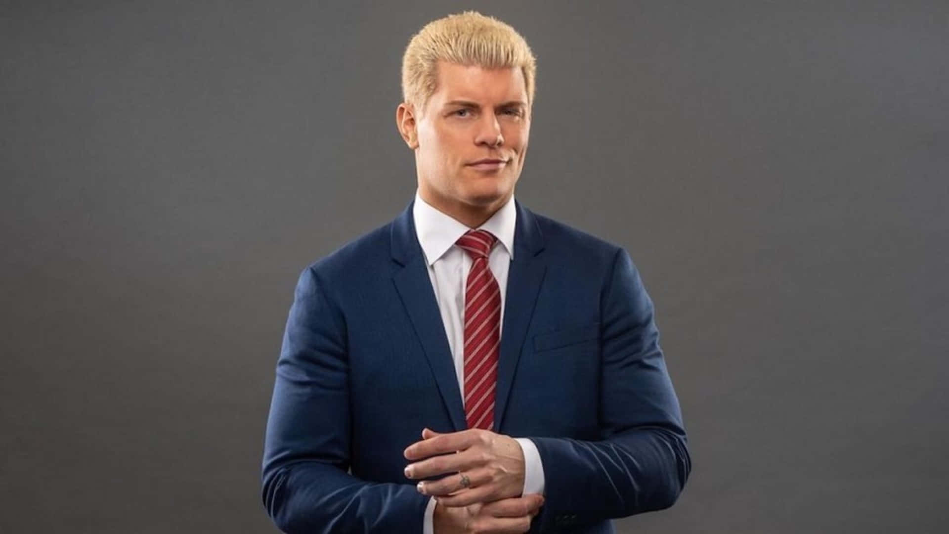 WWE 桌布 壁紙 — 一位自信的男人，留著鉑金色頭髮，身穿一套銳利的藍色西裝和紅色條紋領帶，背景是純灰色的攝影棚。