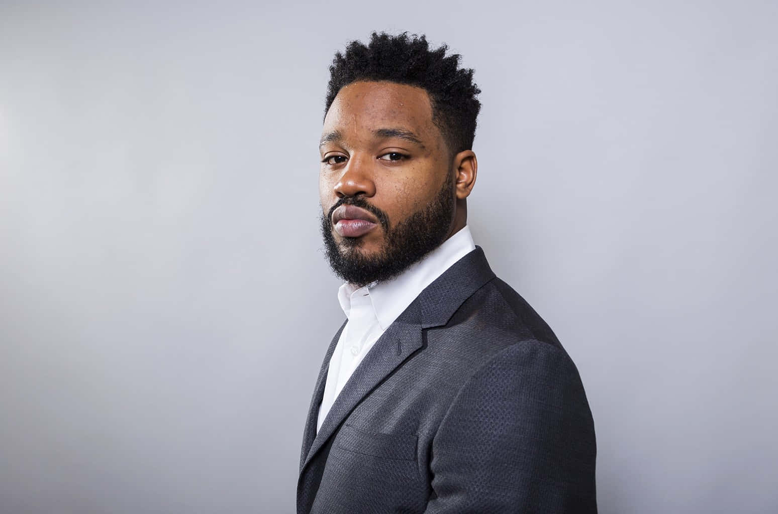 Ryan Coogler 桌布 壁紙 — 一位自信的男人，留著鬍鬚，穿著有質感的西裝，站在純灰色背景前，是專業攝影棚肖像。