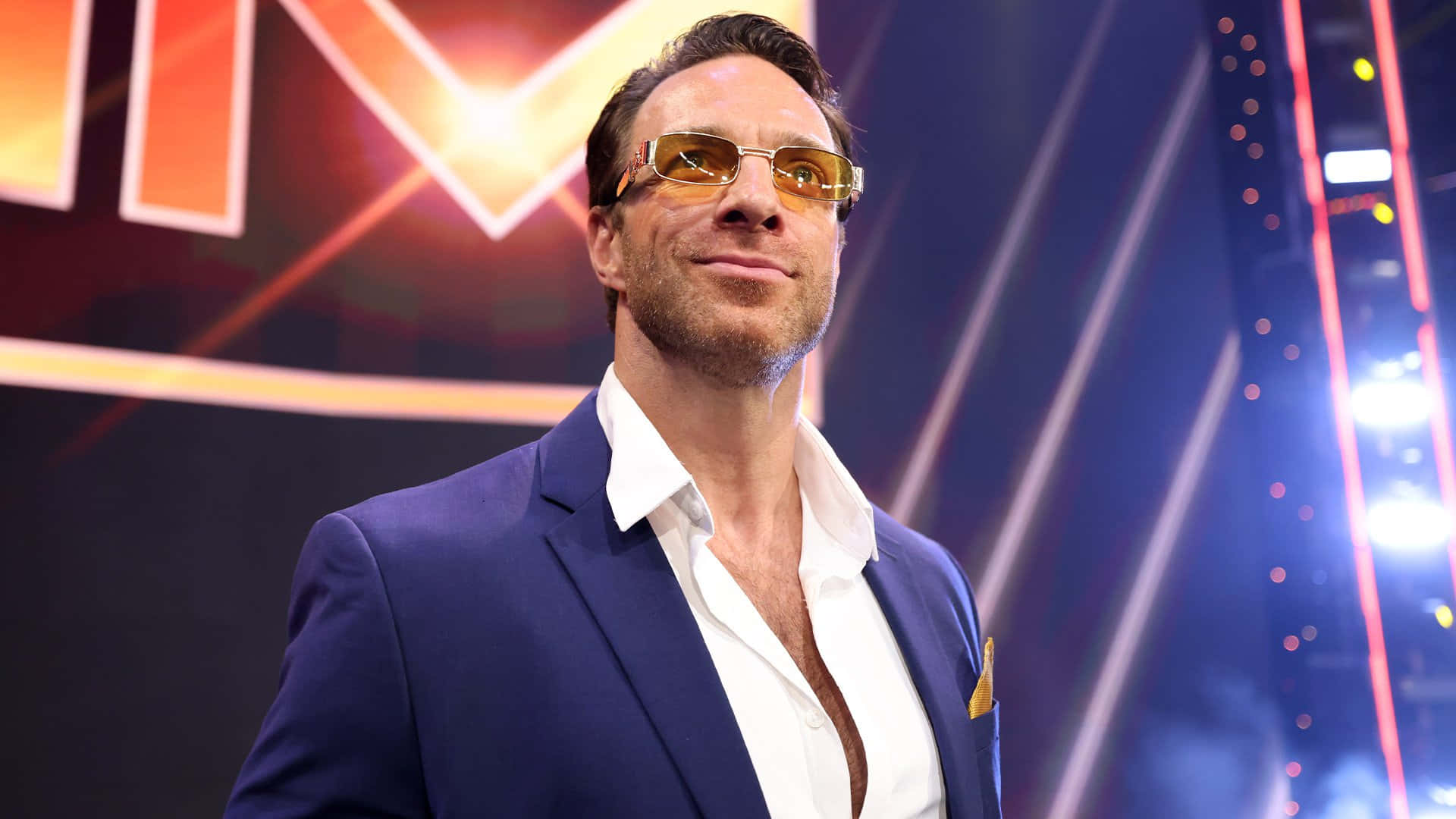 WWE 桌布 壁紙 — 一位自信的男人，身穿藍色西裝和金色太陽眼鏡，站在一個充滿活力的舞台前，舞台上有發光的燈光和一個大的「WWE」標誌。