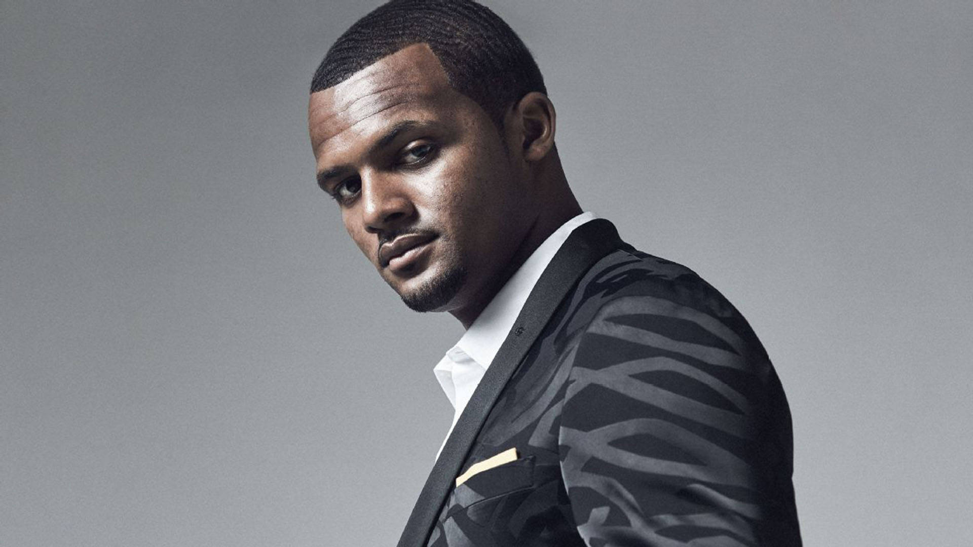 Deshaun Watson 桌布 壁紙 — 一位自信的男人穿著圖案西裝，在純灰色背景前擺姿勢，眼神強烈。