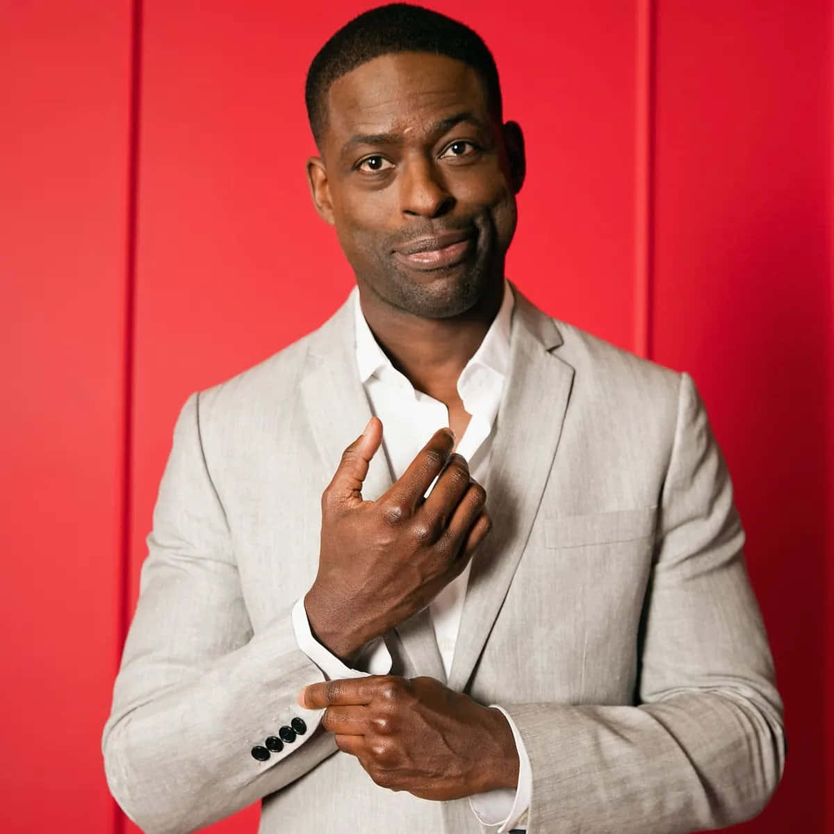 Sterling K Brown 桌布 壁紙 — 一位自信的男人穿著淺灰色西裝，在搶眼的紅色攝影棚背景前調整袖口。