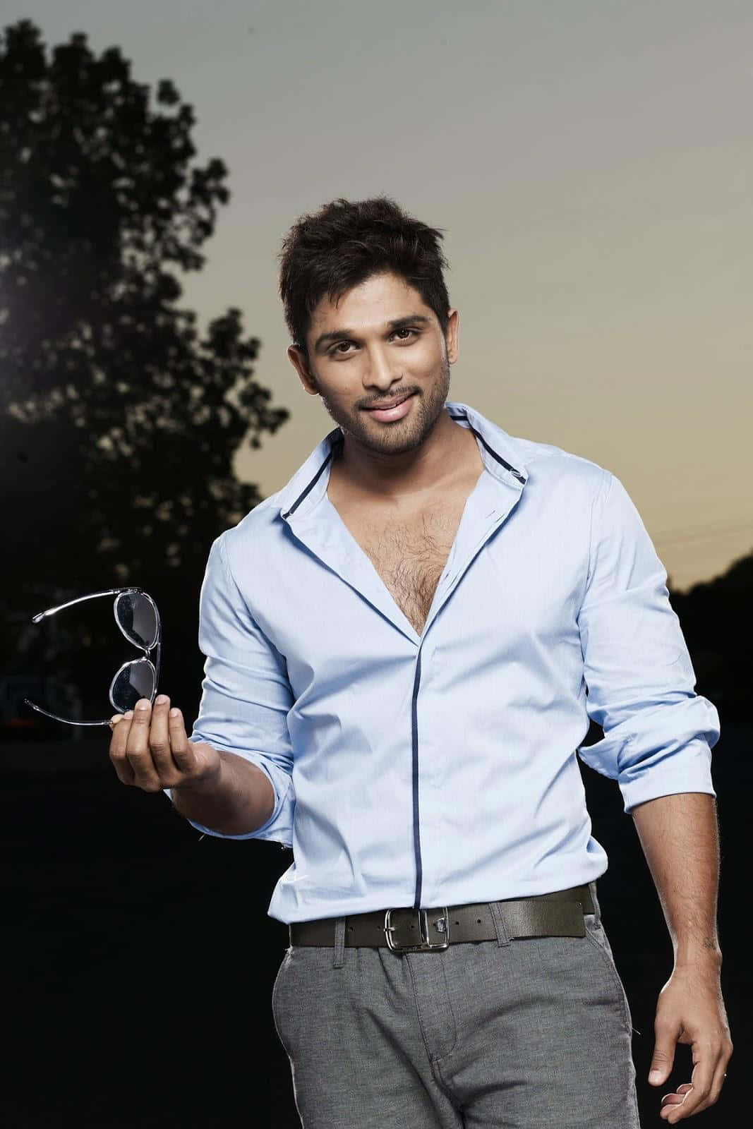 Allu Arjun 桌布 壁紙 — 一位自信的男人穿著淺藍色襯衫，在暮色天空下拿著太陽眼鏡，背景有剪影樹木。