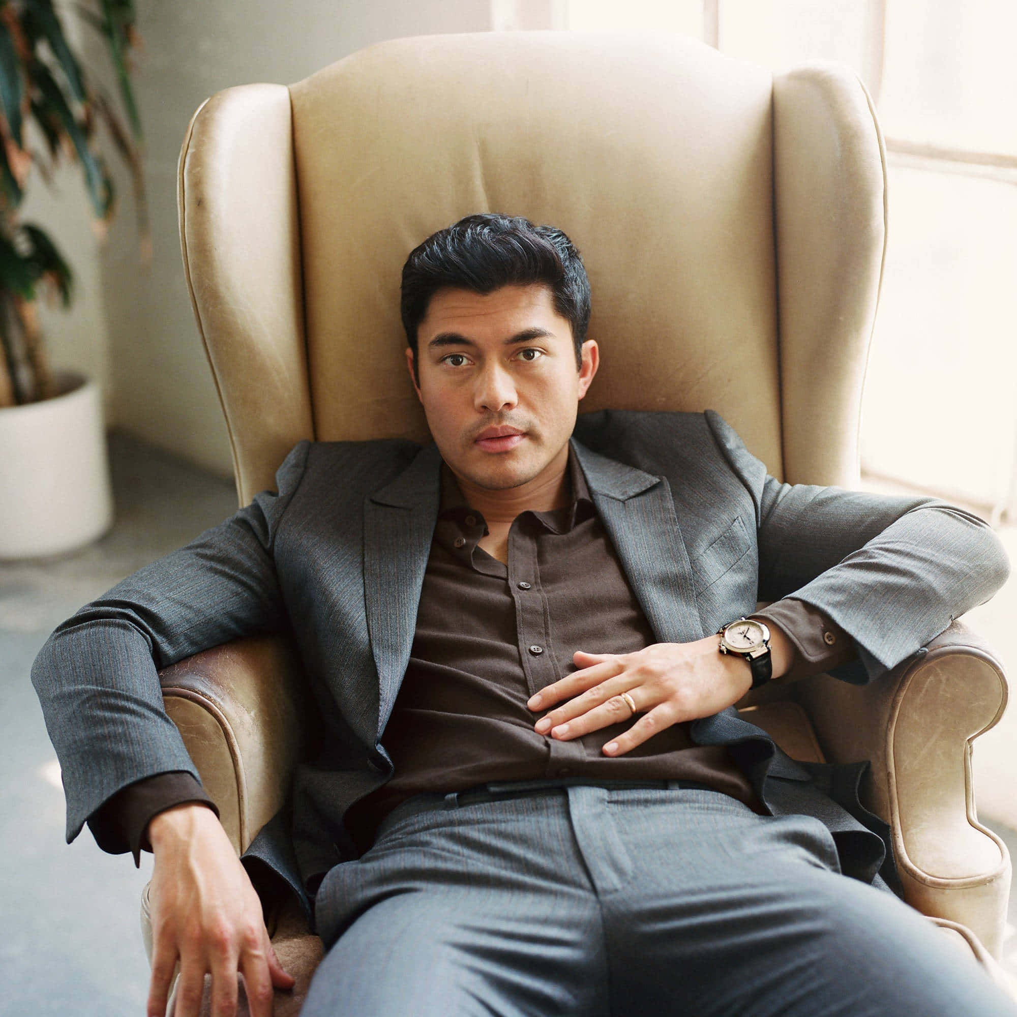 actor 桌布 壁紙 — 一位自信的男人穿著灰色西裝，在米色皮扶手椅上放鬆，背景有盆栽植物。