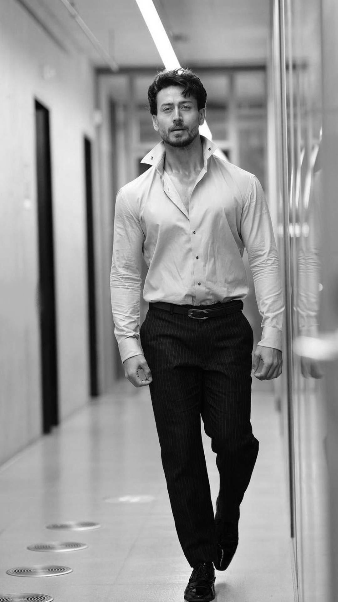 Tiger Shroff 桌布 壁紙 — 一位自信的男人穿著筆挺的襯衫和細條紋褲，在一條時尚現代的走廊上以黑白風格走過。