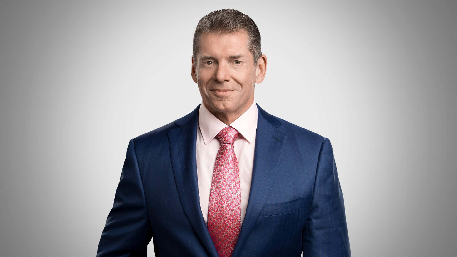 WWE 桌布 壁紙 — 一位自信的男士，身穿剪裁俐落的藍色西裝，搭配圖案粉紅色領帶，在灰色中性攝影棚背景前擺姿勢。
