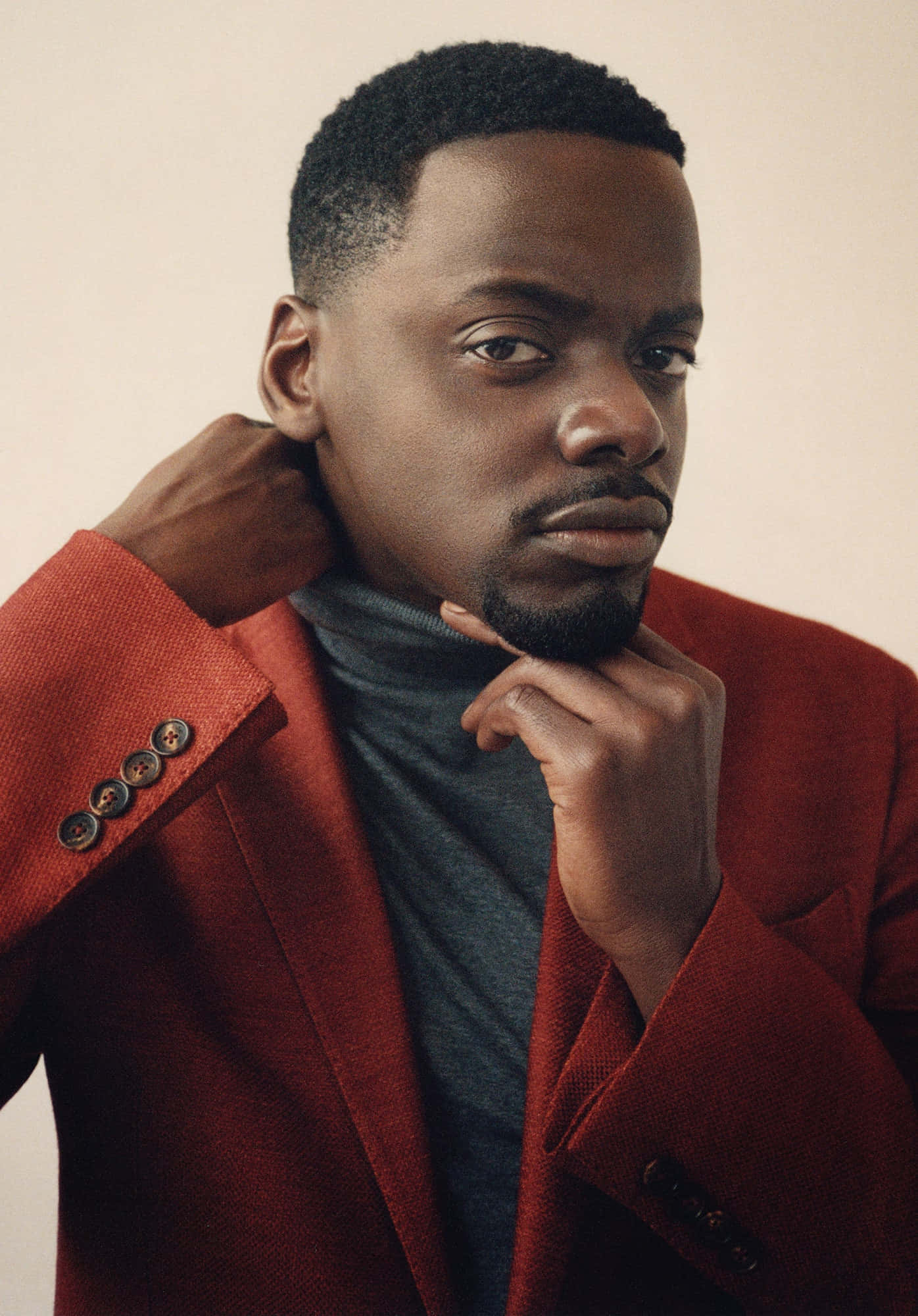 Daniel Kaluuya 桌布 壁紙 — 一位自信的男士穿著大膽的紅色西裝外套和灰色高領毛衣，在柔和的米色背景前將手靠近頸部擺姿勢。