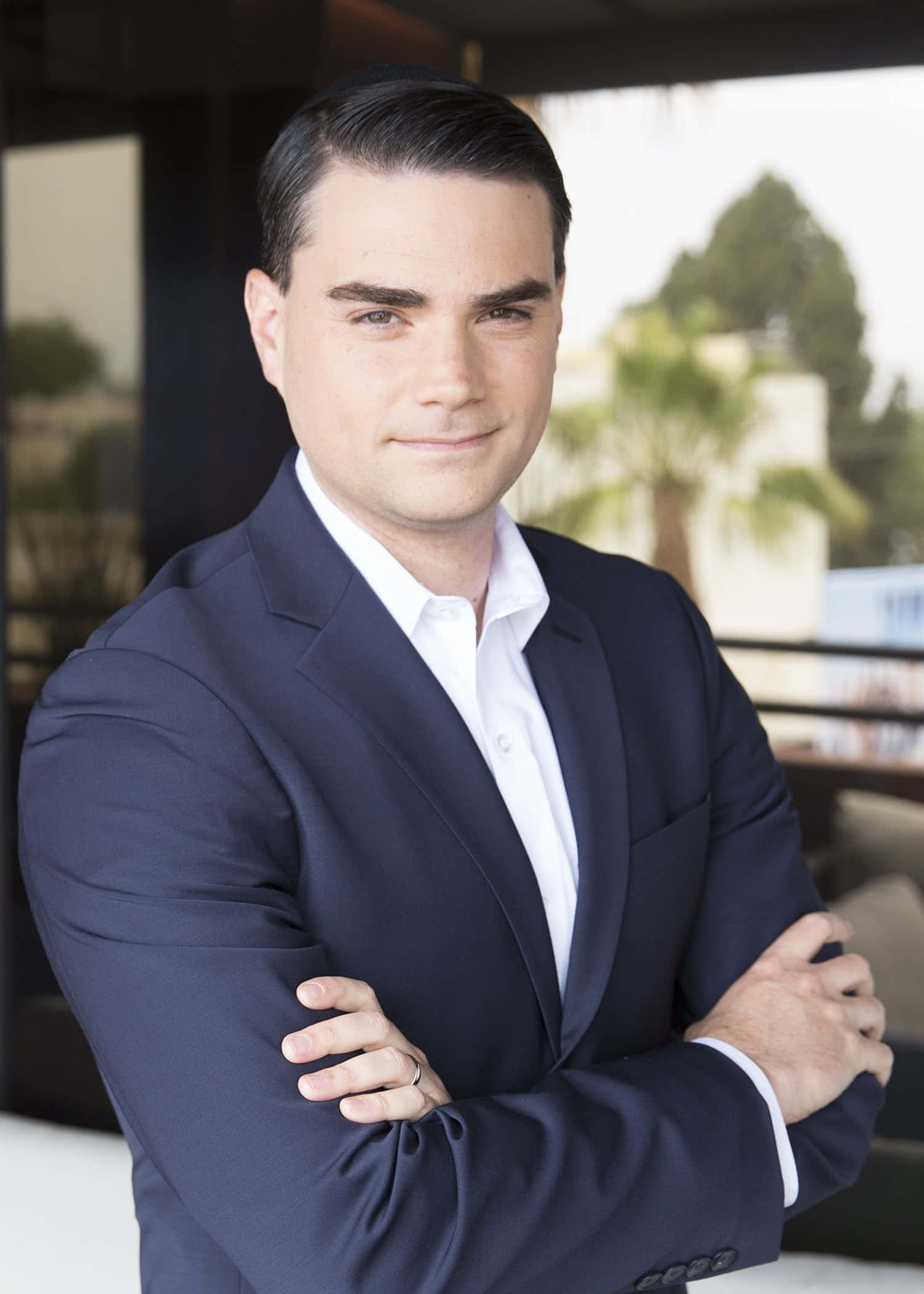 Ben Shapiro 桌布 壁紙 — 一位自信的男士穿著海軍藍西裝，雙手交叉，以現代建築和棕櫚樹為背景站著。