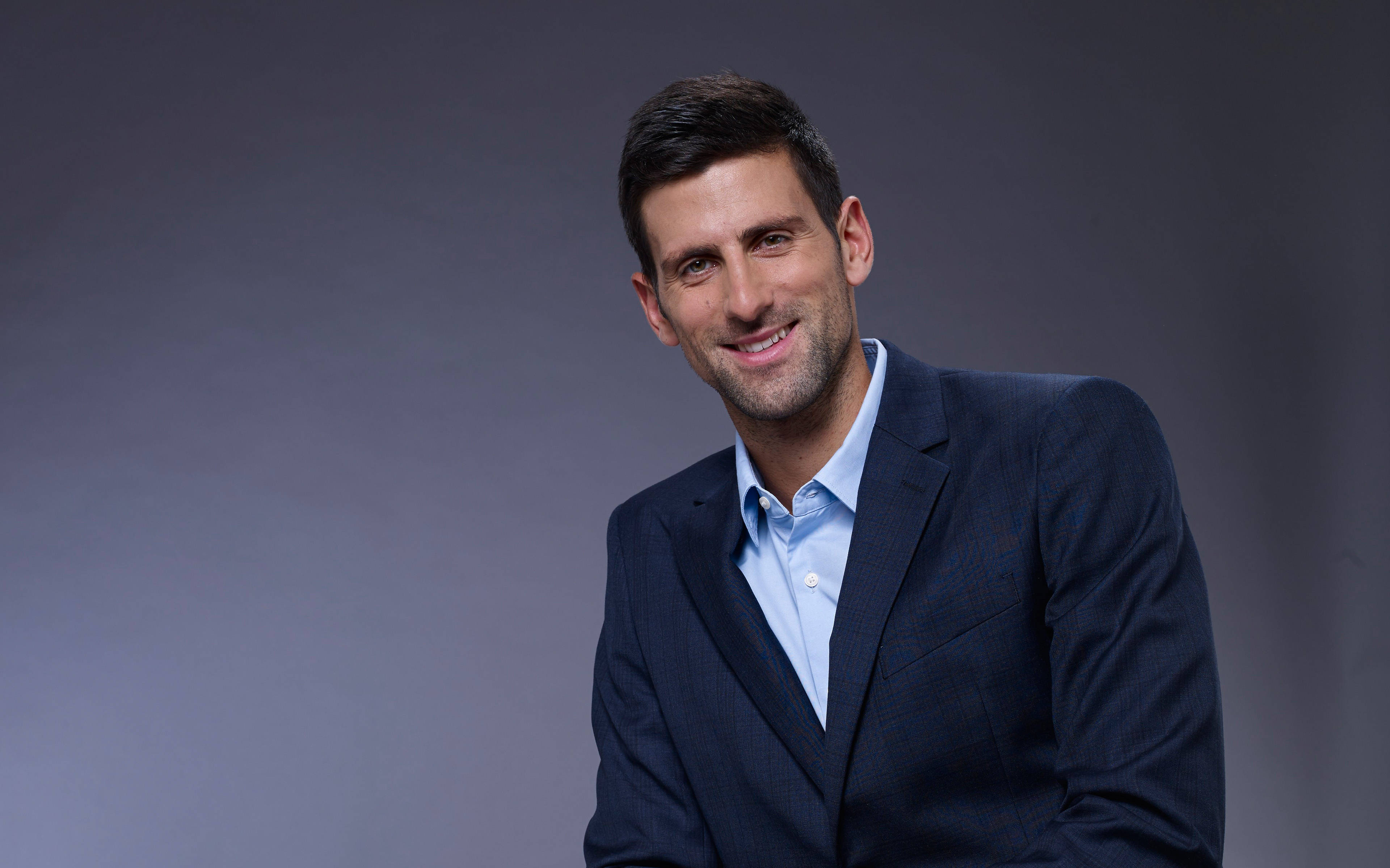 Novak 桌布 壁紙 — 一位自信的男士穿著深色西裝和淺藍色襯衫，在柔和的灰色攝影棚背景下溫暖微笑。
