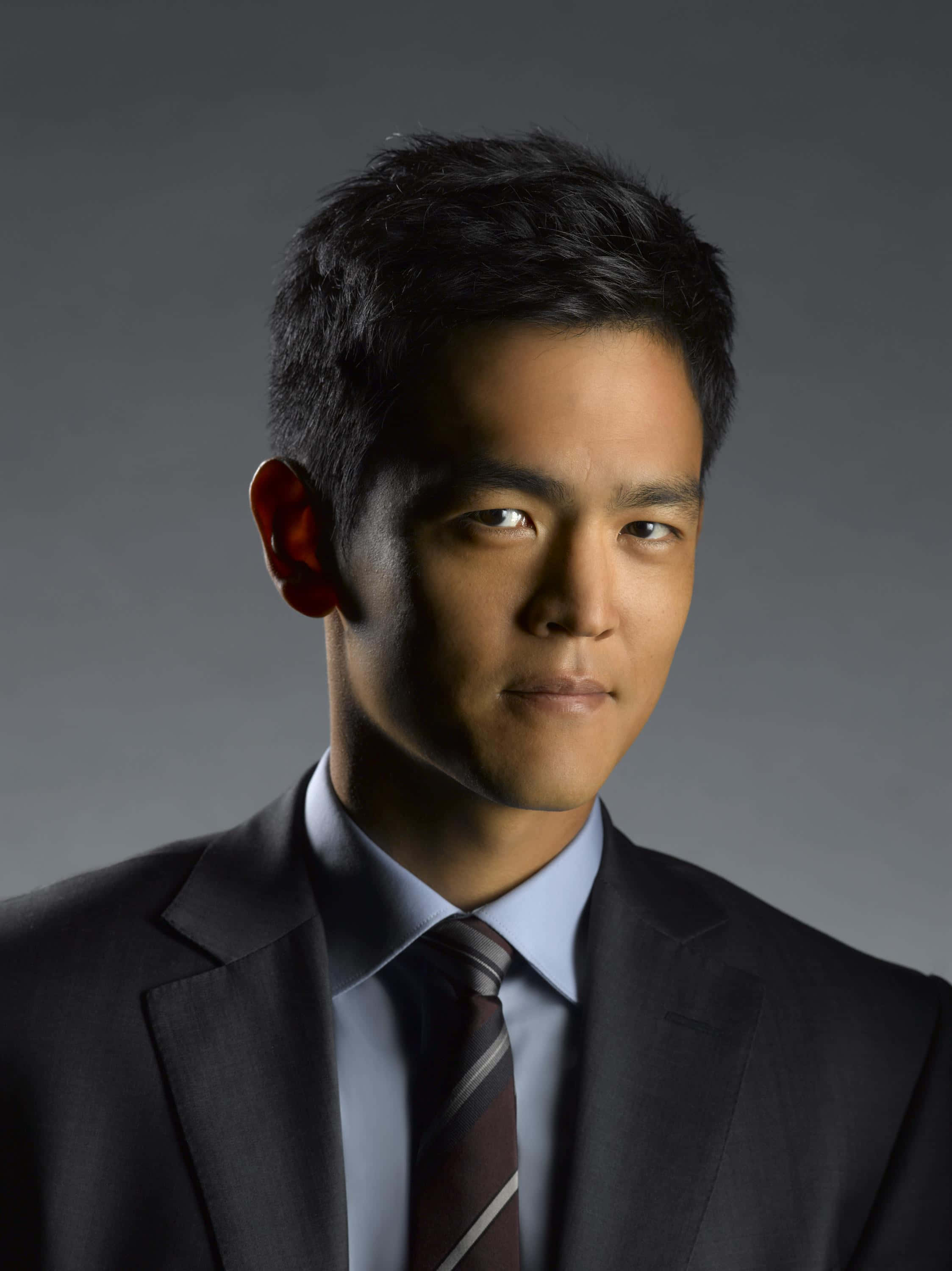 John Cho 桌布 壁紙 — 一位自信的男子身穿深色西裝和條紋領帶，在柔和的灰色攝影棚背景前擺姿勢，光線柔和。