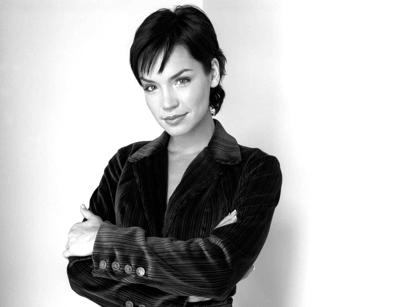 Ashley Scott 桌布 壁紙 — 一位自信的短黑髮女性，靠著白牆站立，身穿質感黑色西裝外套，雙臂交叉。