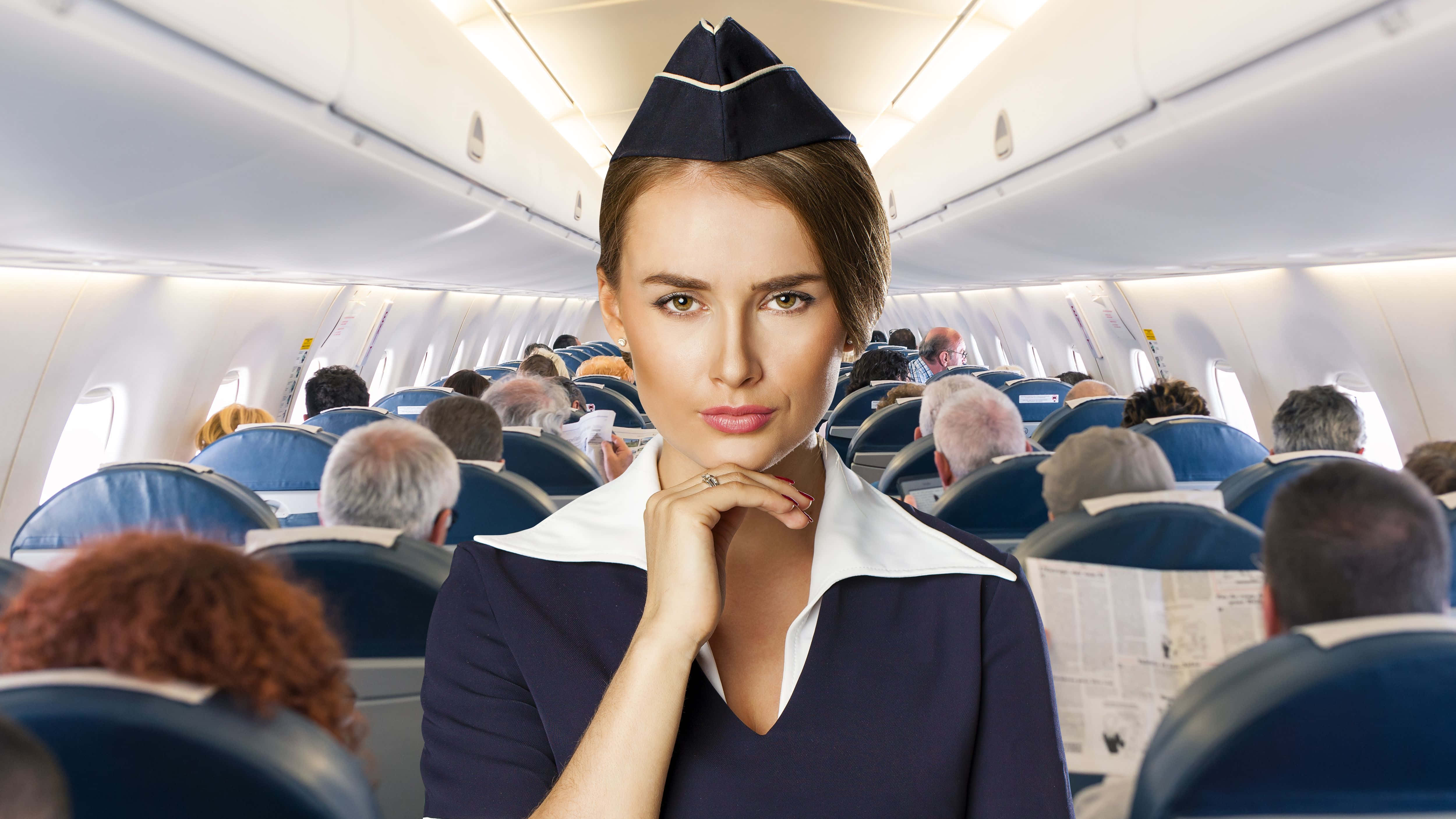 Flight Attendant 桌布 壁紙 — 一位自信的空中服務員穿著制服，坐在光線明亮的機艙乘客中，直視鏡頭。