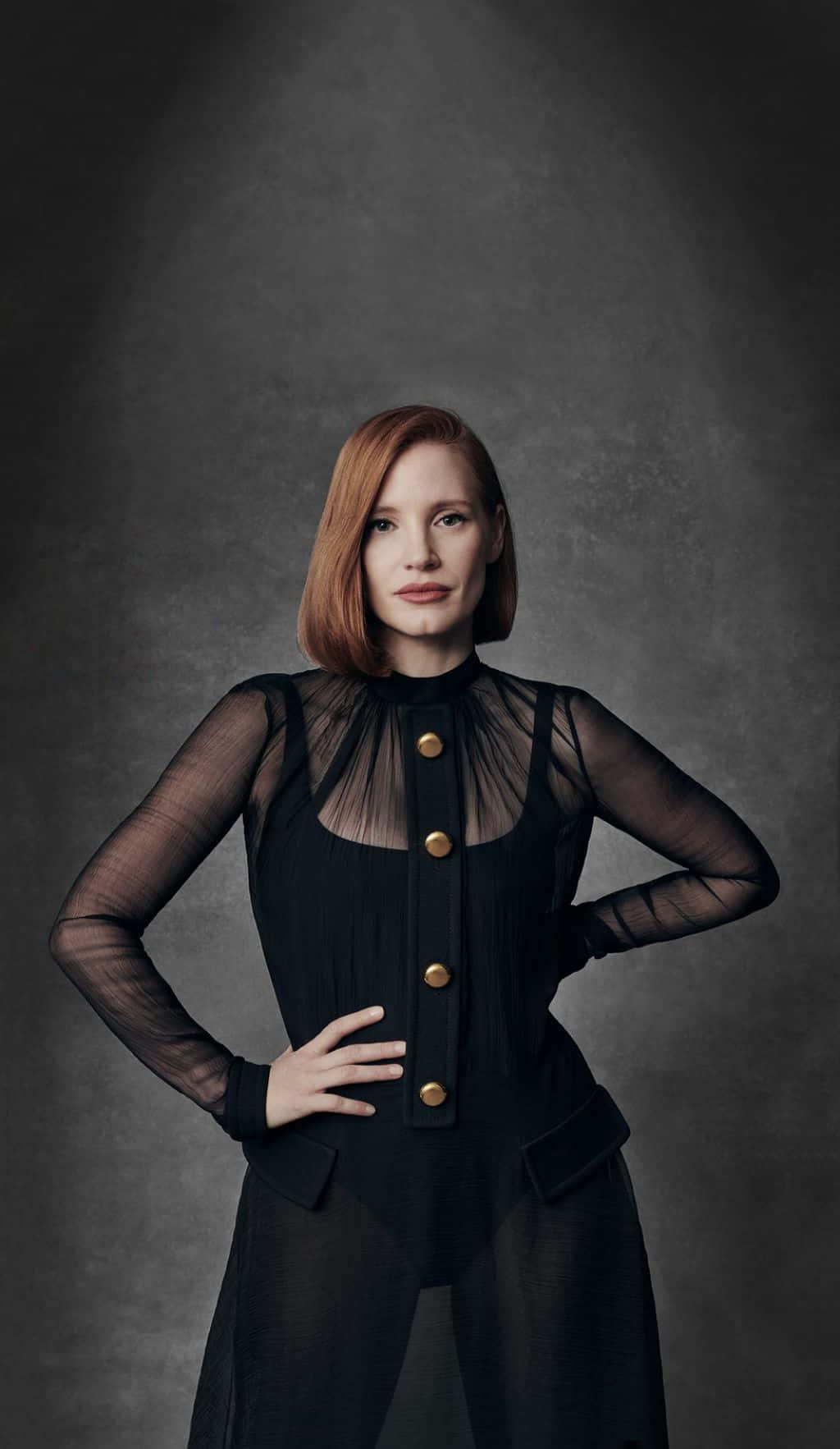 jessica chastain 桌布 壁紙 — 一位自信的紅髮女性，身穿黑色透明袖服裝，搭配金色鈕扣，站在紋理灰色背景前。