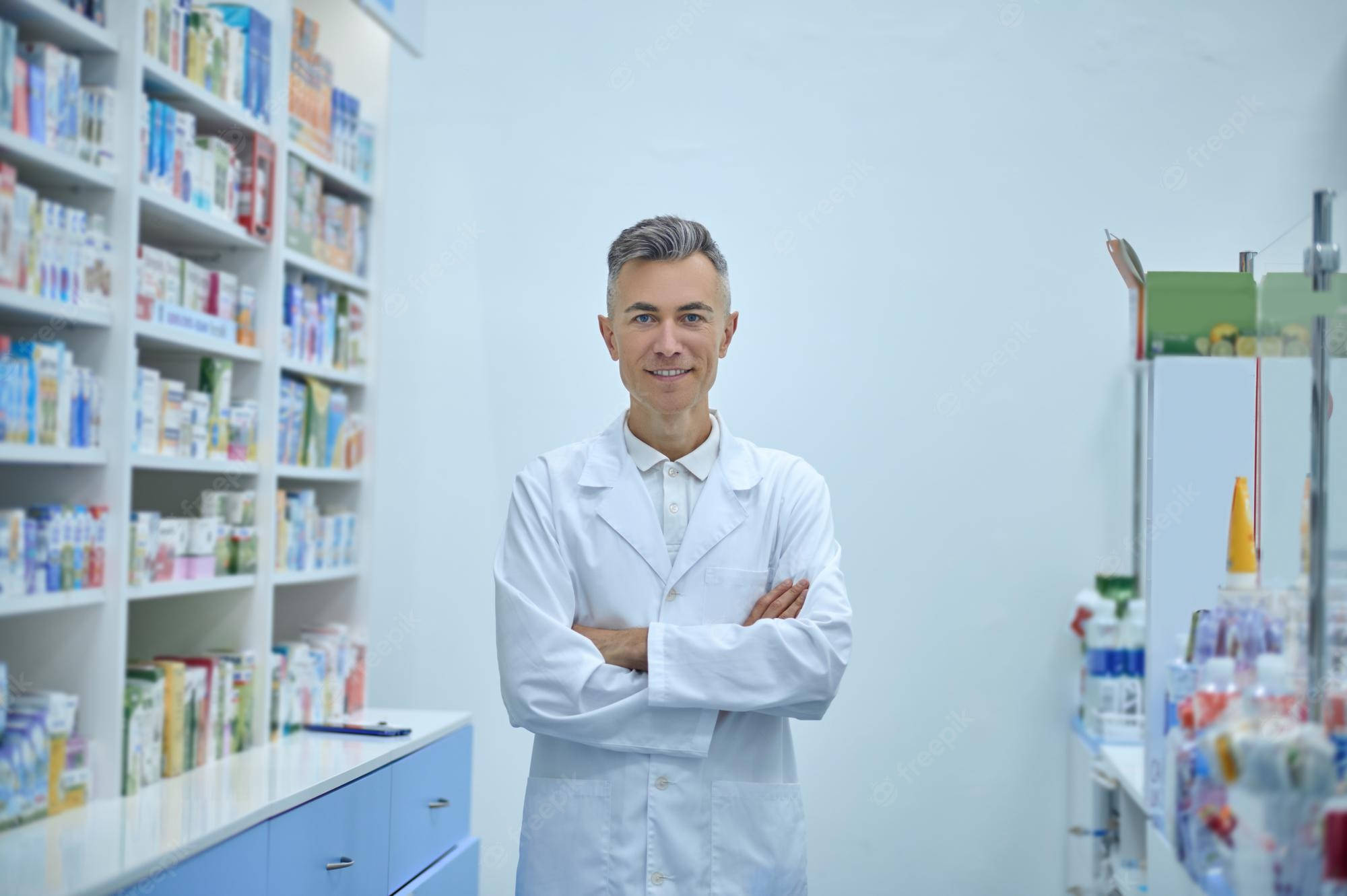 professional 桌布 壁紙 — 一位自信的藥劑師身穿白袍，在光線充足的藥局走道中交叉雙臂站立，周圍是色彩繽紛的藥品貨架。