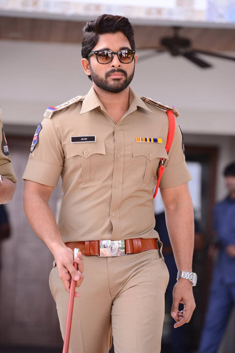 Allu Arjun 桌布 壁紙 — 一位自信的警察穿著卡其色制服和太陽眼鏡，在明亮的日光下手持警棍向前邁步。