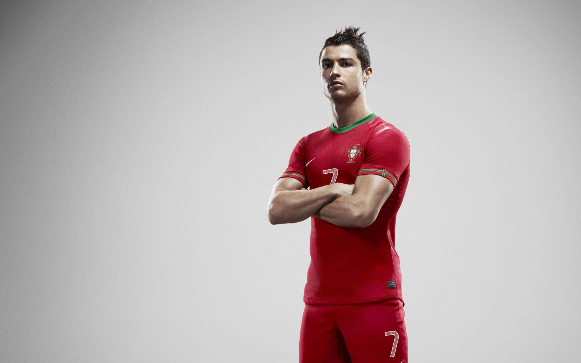 Cr7 桌布 壁紙 — 一位自信的足球員身穿紅色葡萄牙球衣，在純灰色攝影棚背景前交叉雙臂站立。